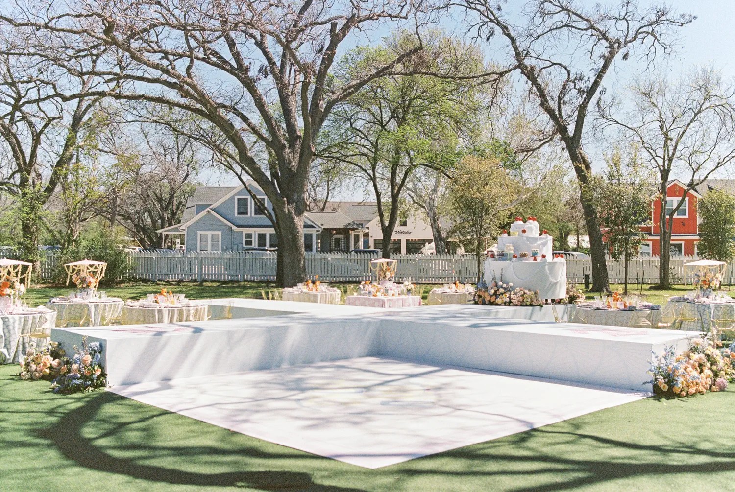 Outdoor brunch wedding reception lawn with white dance floor, giant tiered cake display topped with red cherries, and pastel tables