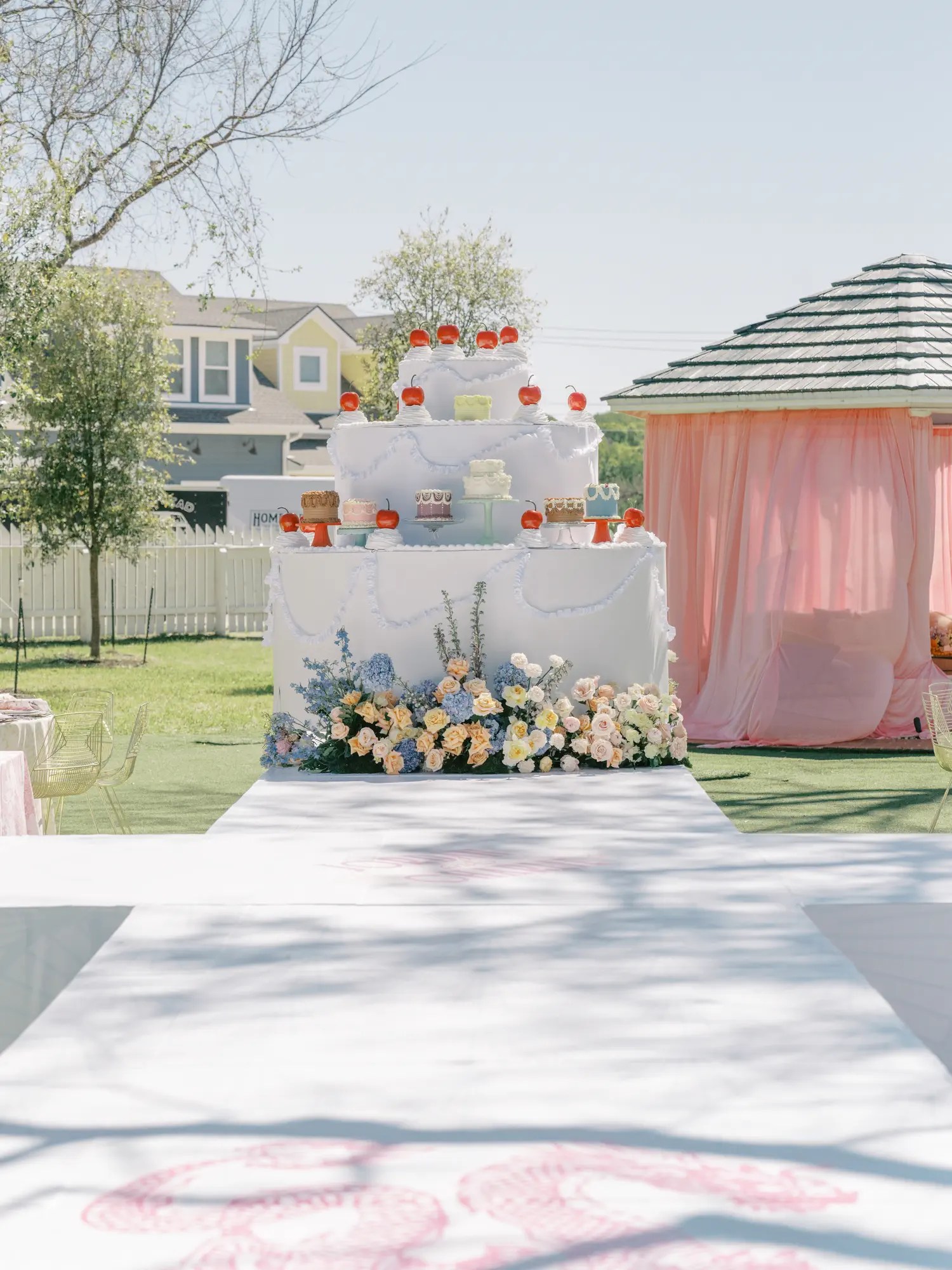 Giant pale blue tiered wedding cake display with white ruffled swags, oversized red cherries, and small cakes surrounded by roses and hydrangea