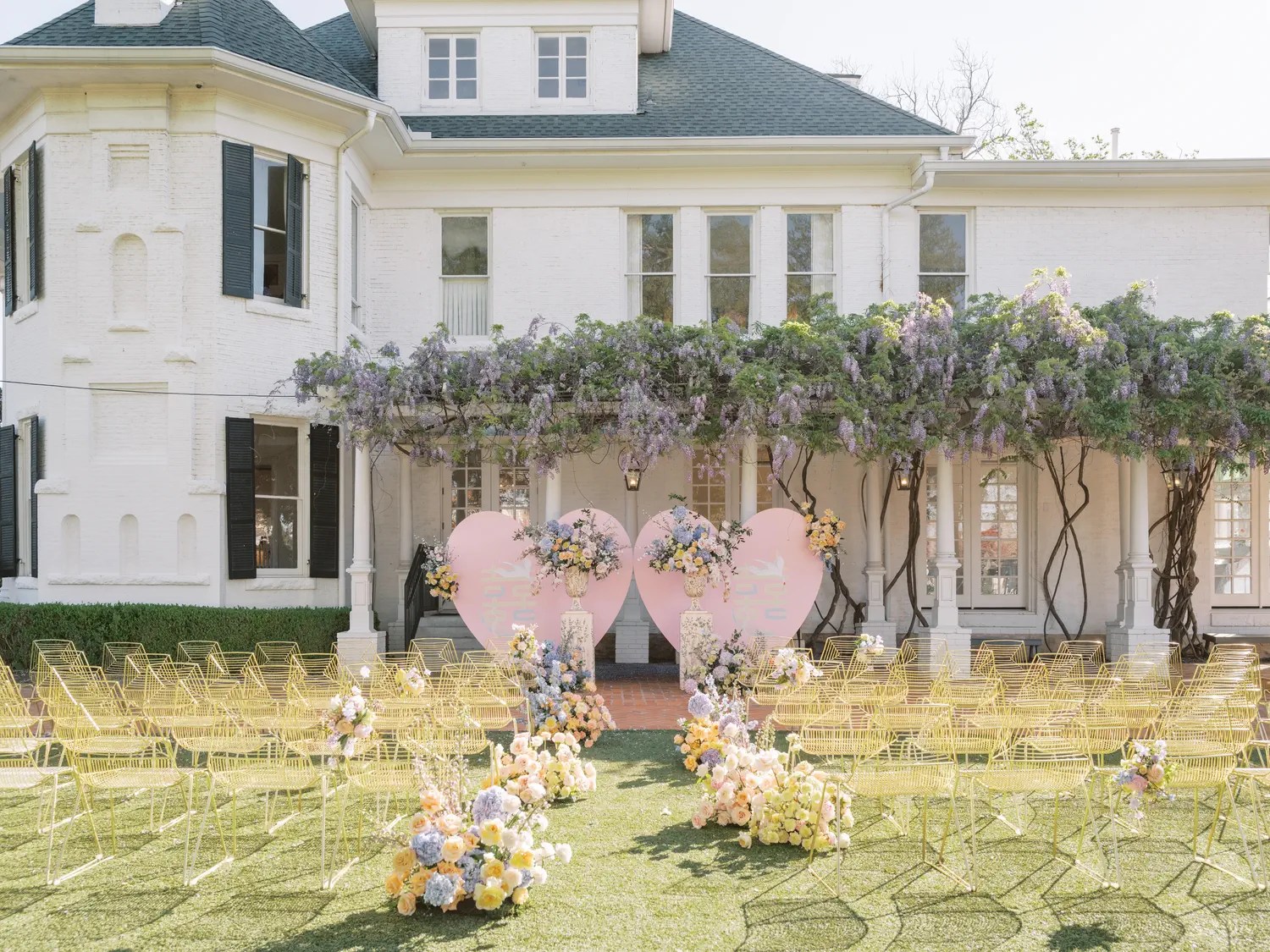 Outdoor spring wedding ceremony at Woodbine Mansion in Austin with yellow wire chairs, grass aisle, pink heart backdrop, and wisteria-covered porch