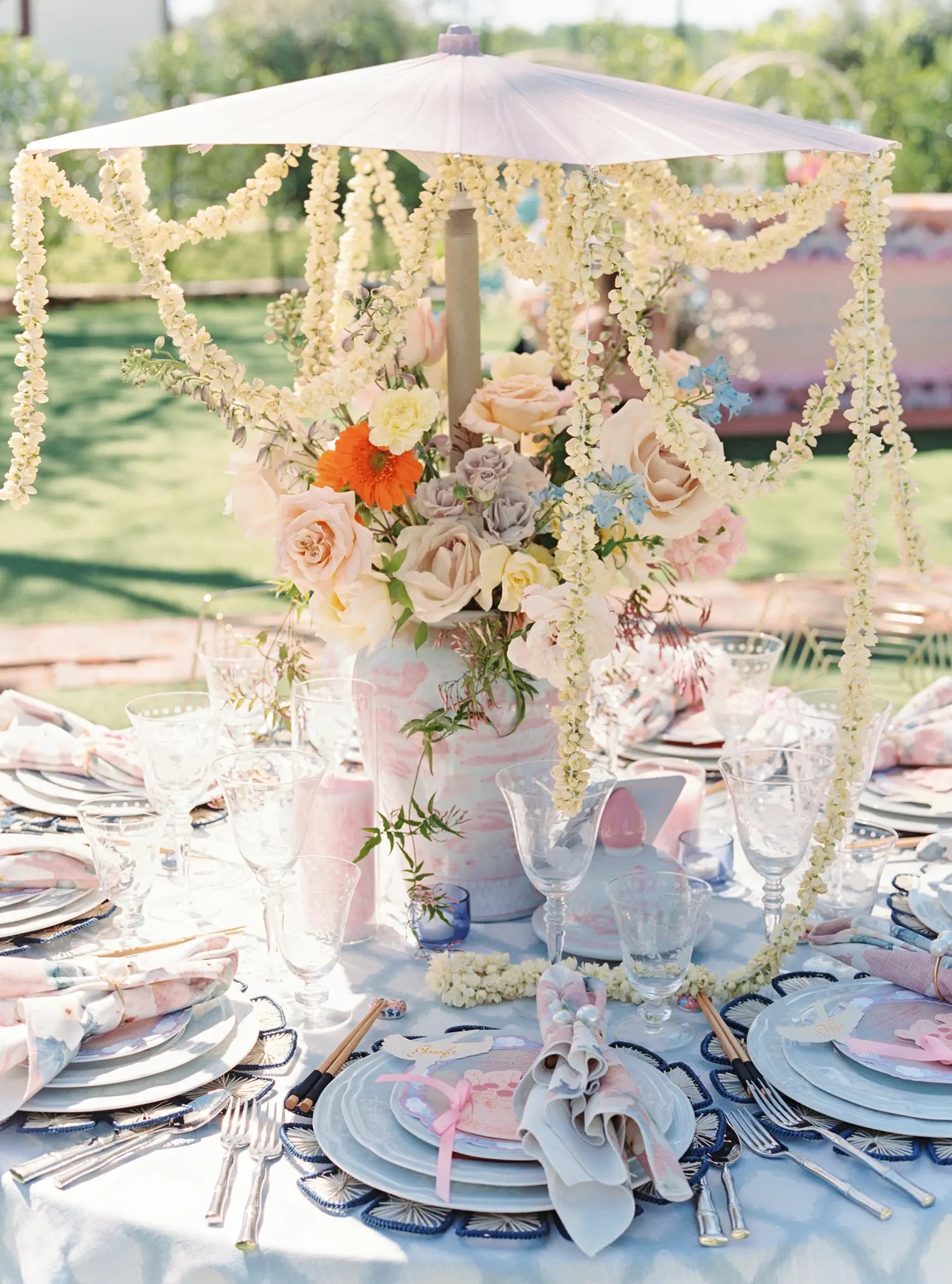 Brunch reception table with pale blue linen, navy shell chargers, pastel garden rose centerpiece, and cream flower garland hung from a lilac parasol