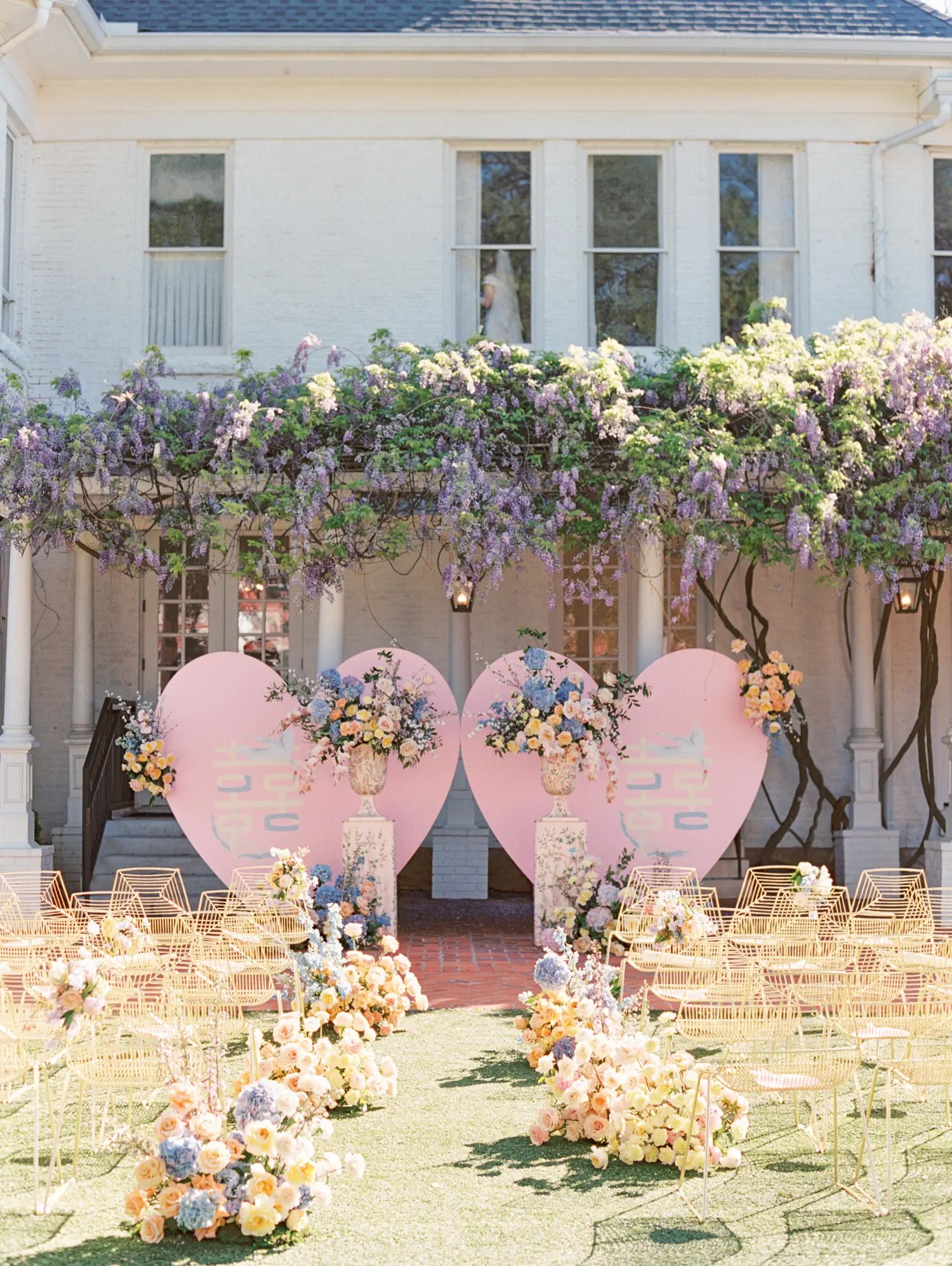 Pastel flowers lining a grass ceremony aisle toward pink double happiness hearts in front of a white brick mansion in Austin, Texas