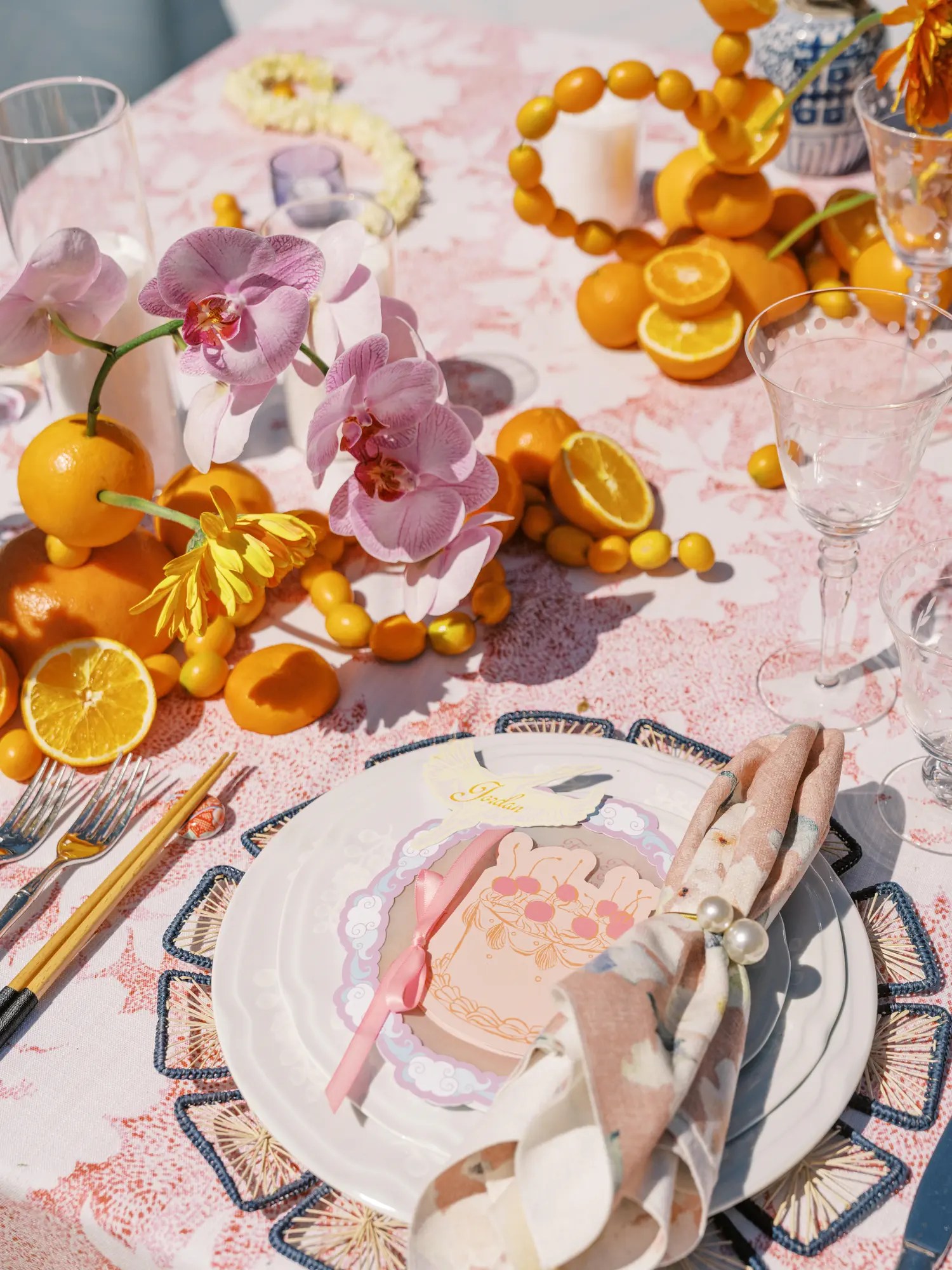 Place setting on a pink floral tablecloth scattered with oranges, kumquats, and pink orchids, set with chopsticks and forks