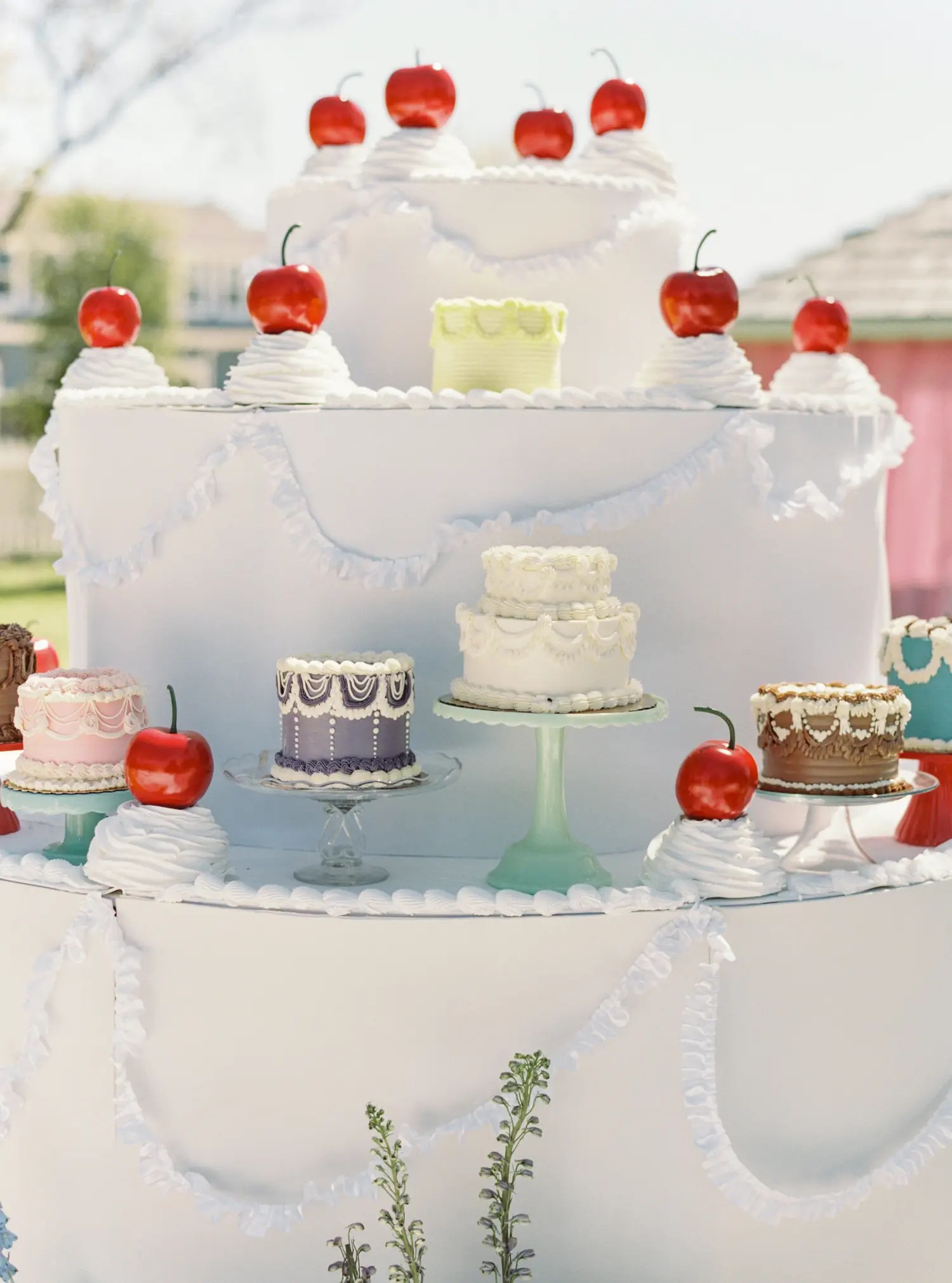 Close-up of giant cake display tiers holding pink, lavender, chocolate, and teal mini cakes with oversized red cherries