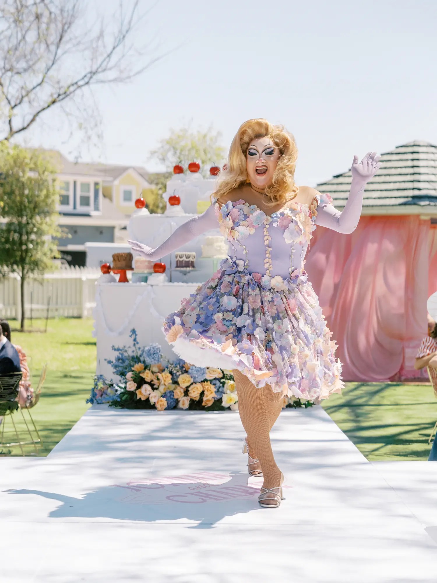 Drag performer in pastel 3D floral off-the-shoulder dress and lavender gloves dancing in front of a giant tiered cake display