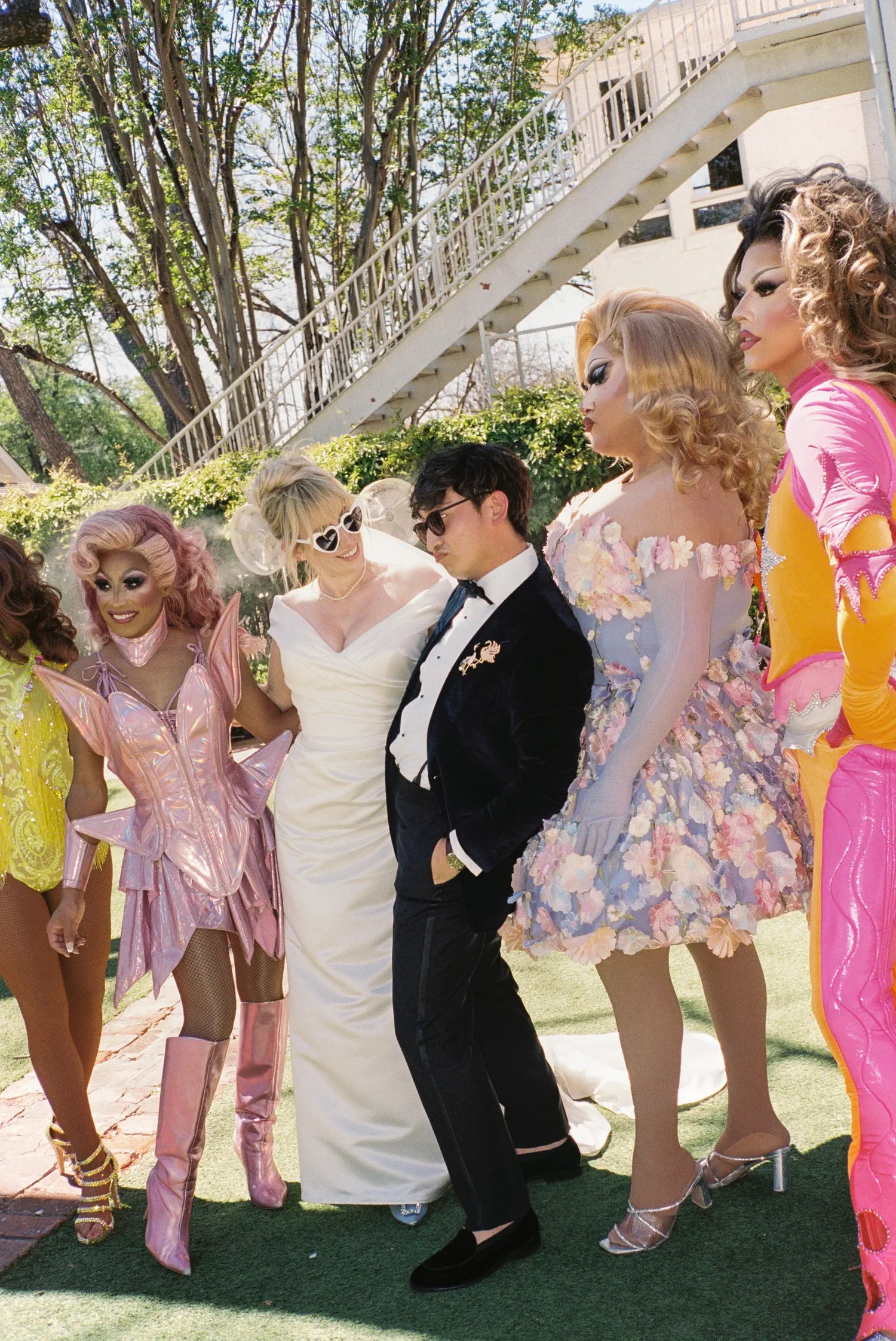 Bride in white heart-shaped sunglasses and groom in velvet tuxedo with four Extragrams drag queens in costume on the lawn
