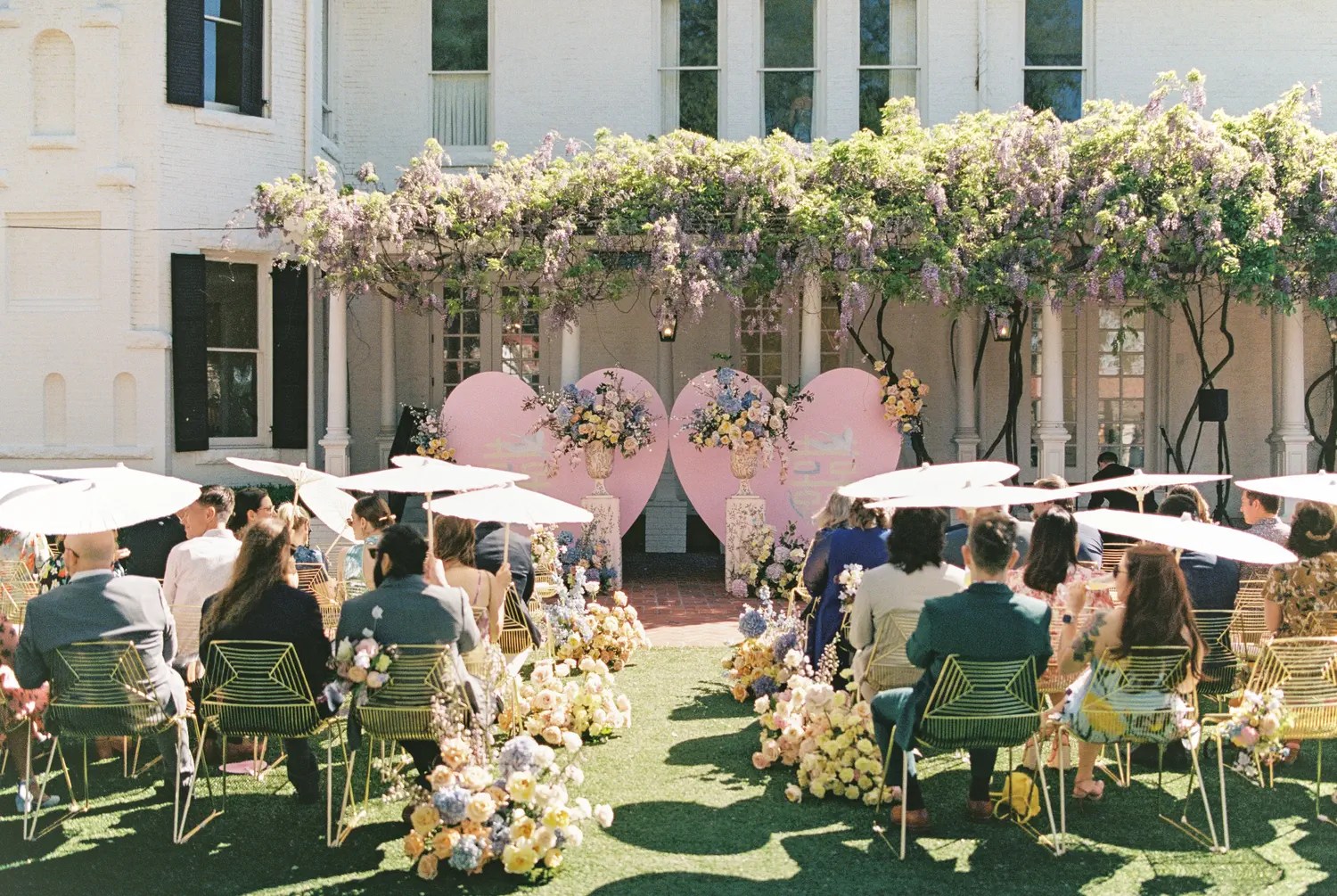 Guests under white parasols at an outdoor daytime wedding ceremony with pink heart altar and wisteria pergola