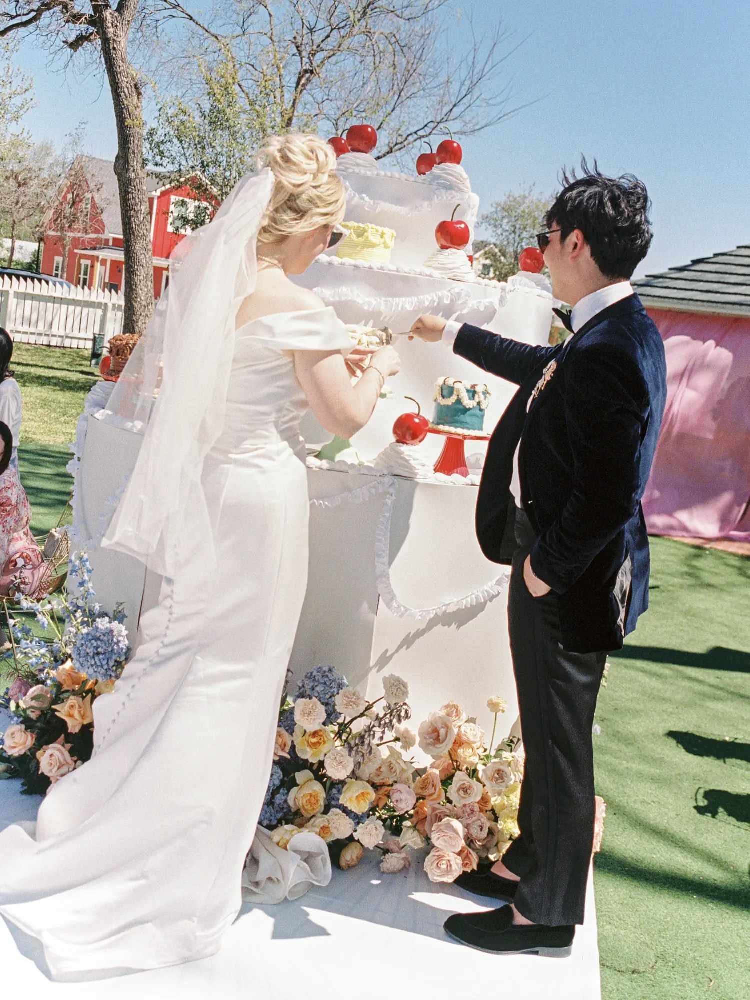 Bride in satin gown and cathedral veil and groom in navy velvet suit cutting a giant tiered cake topped with red cherries