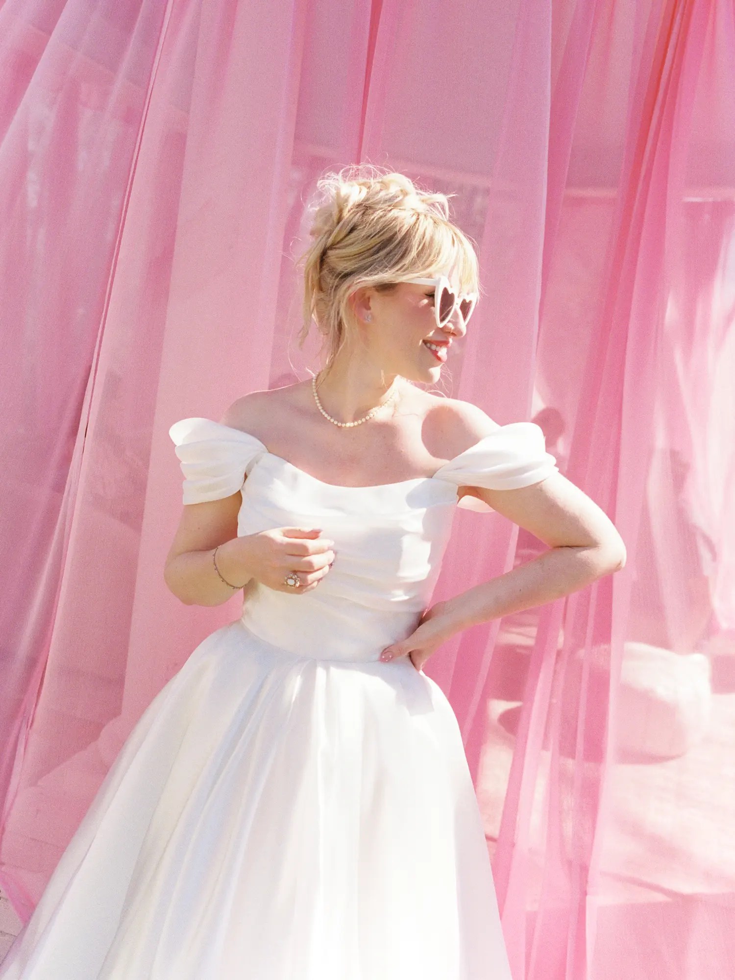Bride in off-the-shoulder satin puff-sleeve dress, pearl necklace, and white heart-shaped sunglasses against sheer pink drapery