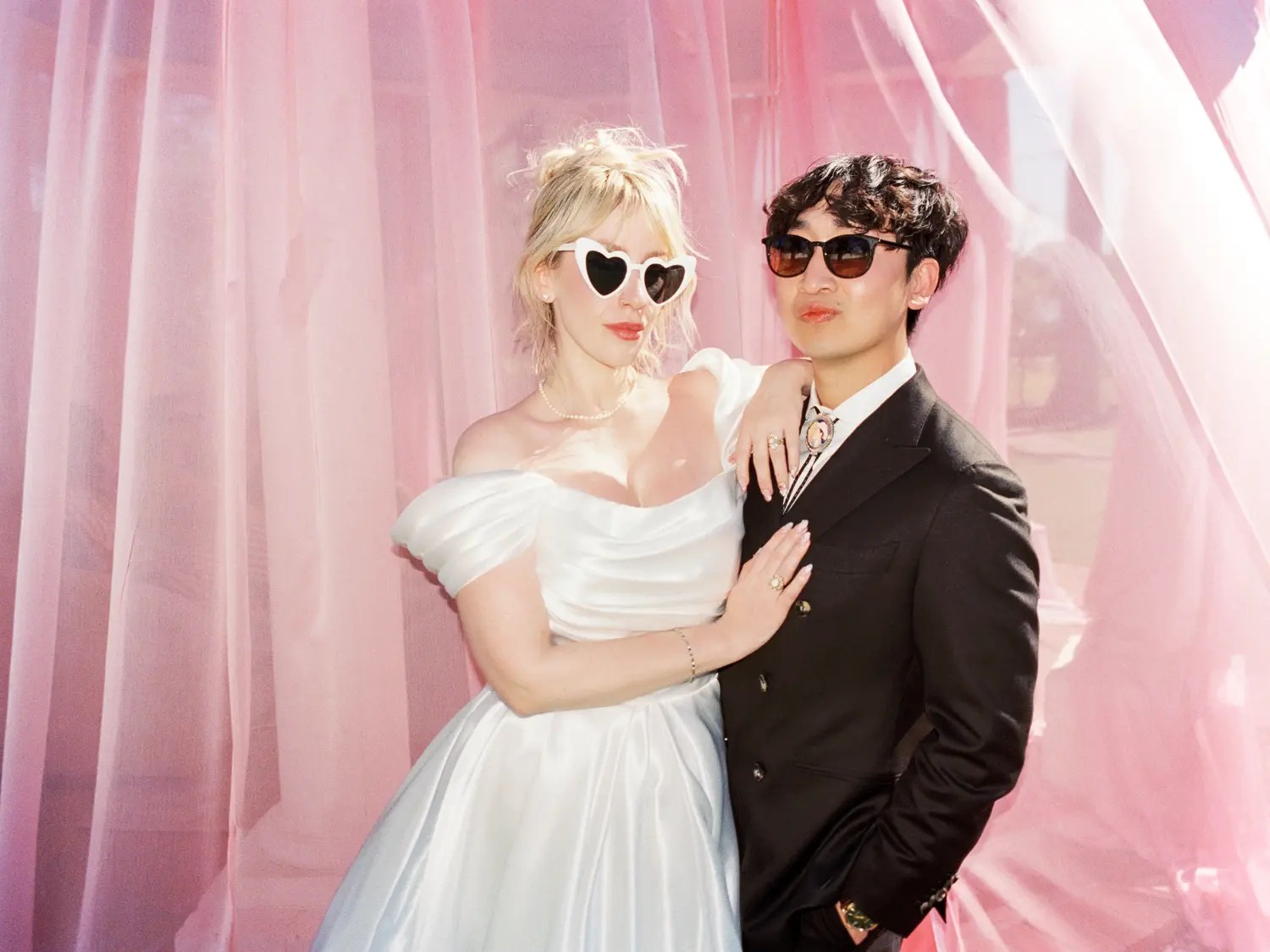Couple in sunglasses, bride in satin puff-sleeve dress and groom in double-breasted suit with bolo tie, in front of sheer pink curtains