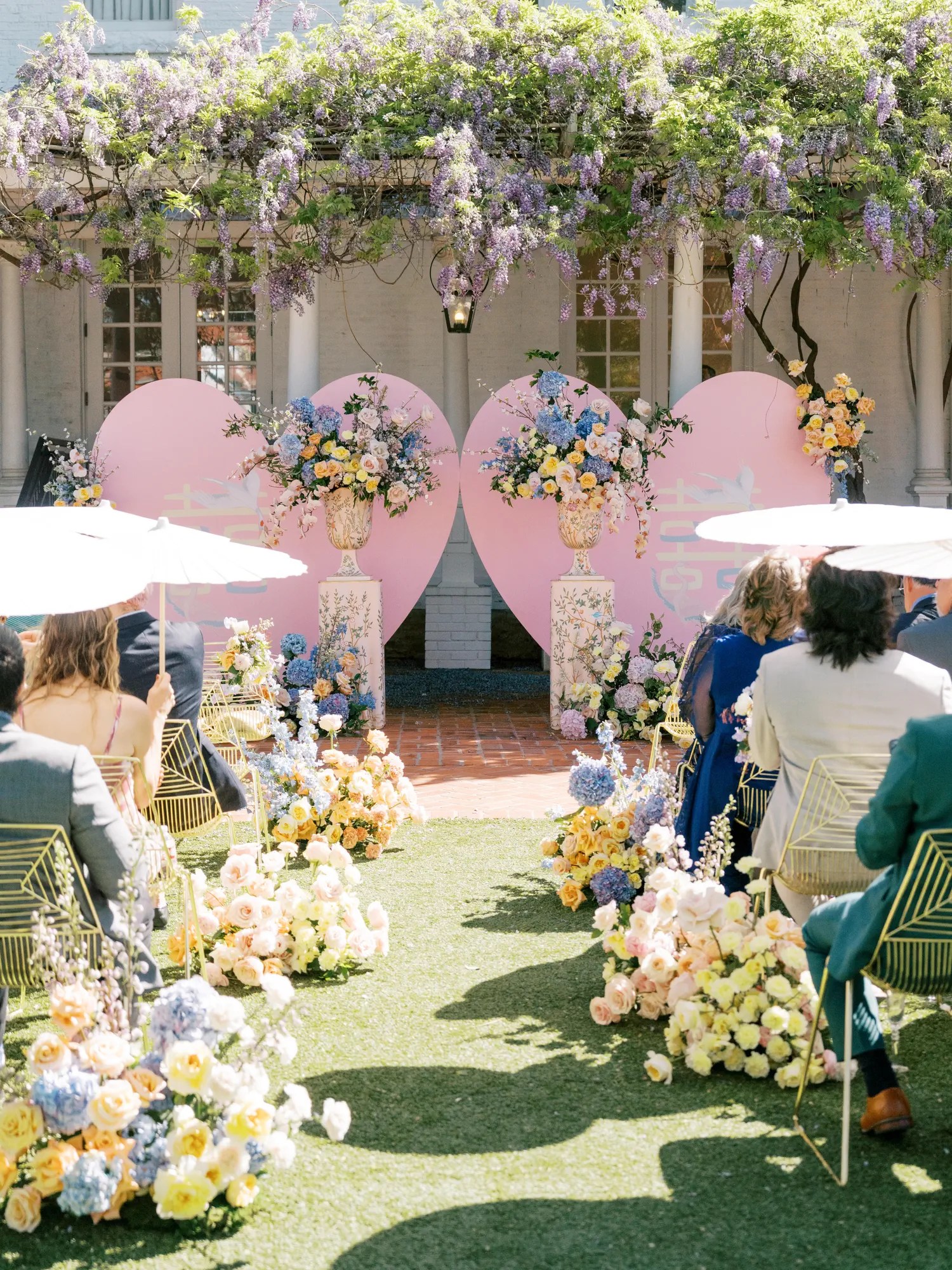 Ceremony aisle lined with peach, blush, and blue blooms on the grass leading to a pink double happiness heart altar under wisteria