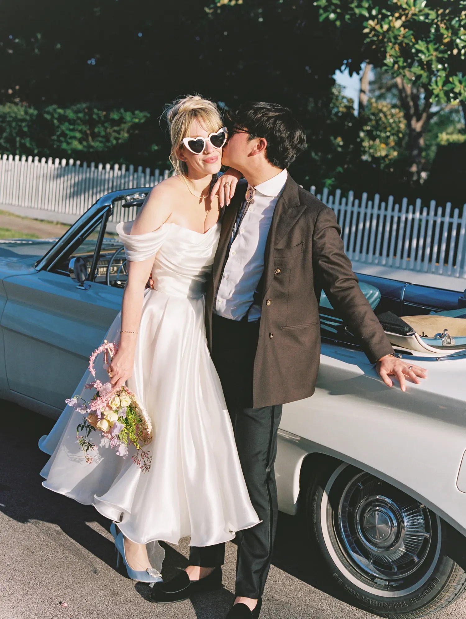 Bride in Olivia Bottega tea-length satin dress with blue kitten heels and groom in brown blazer and bolo tie beside a pale blue vintage convertible from Comal Classics
