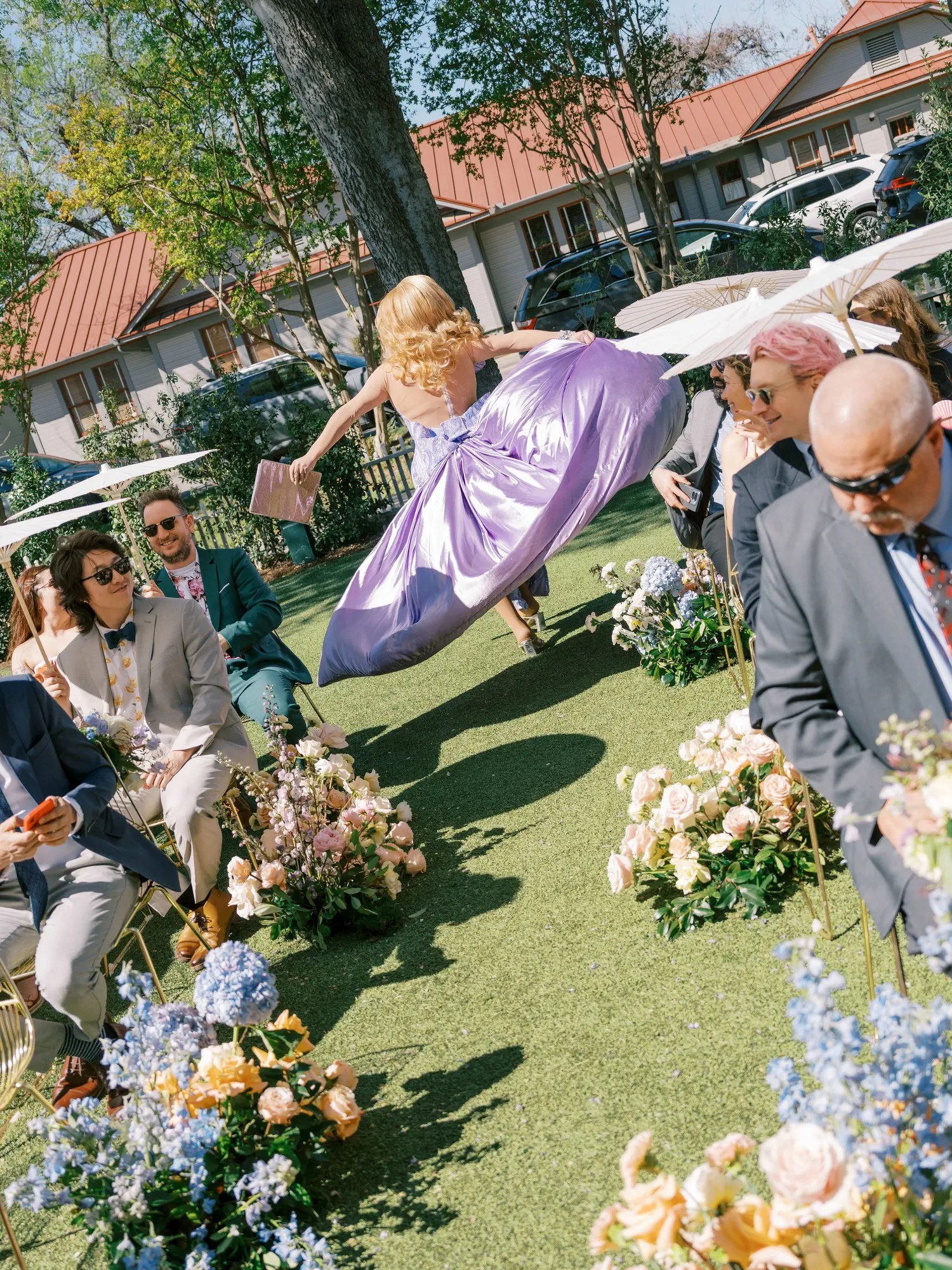 Drag queen officiant in lilac satin gown walking the outdoor ceremony aisle past guests with white parasols