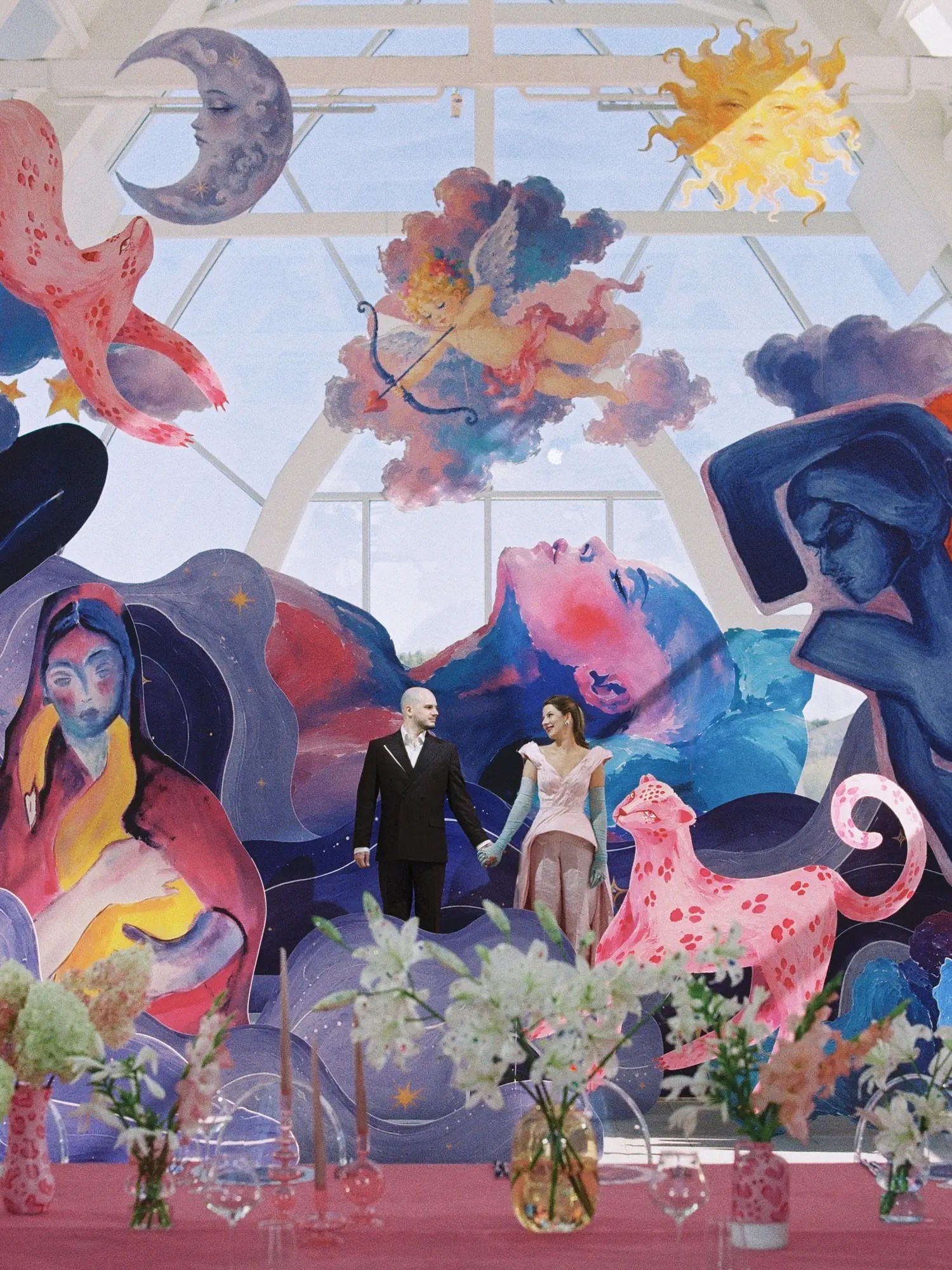 Elena and Alexandr holding hands before a floor-to-ceiling painted mythological mural with a pink leopard in the glass-roofed hall at Soma Island, Voronezh