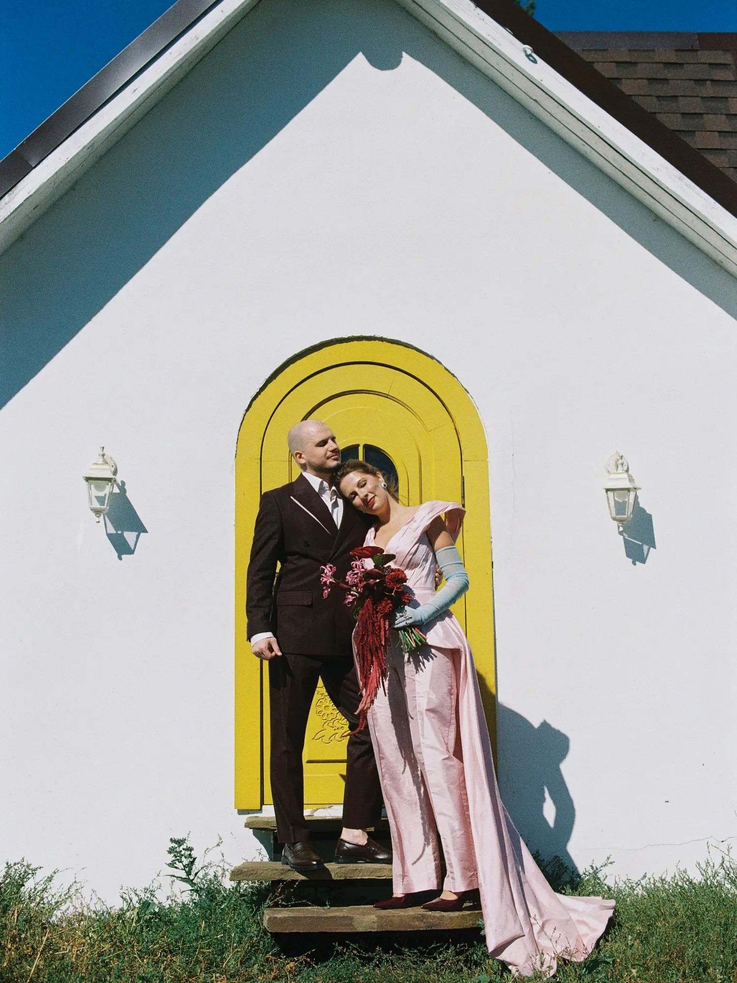 Bride in blush pink off-shoulder gown with pale blue elbow gloves and groom in burgundy suit at a white chapel with a yellow arched door
