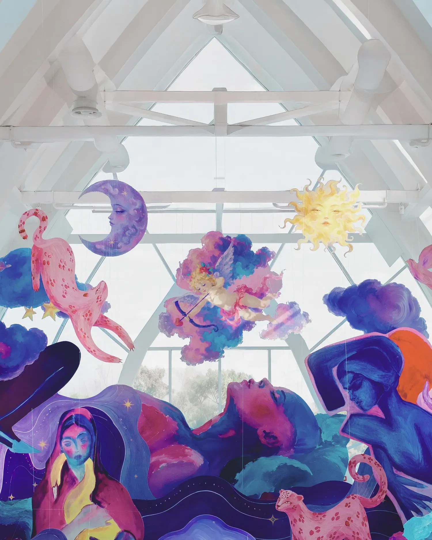 View up into a glass wedding venue ceiling hung with painted cut-outs of a crescent moon, sun, cupid and pink leopard above a mural