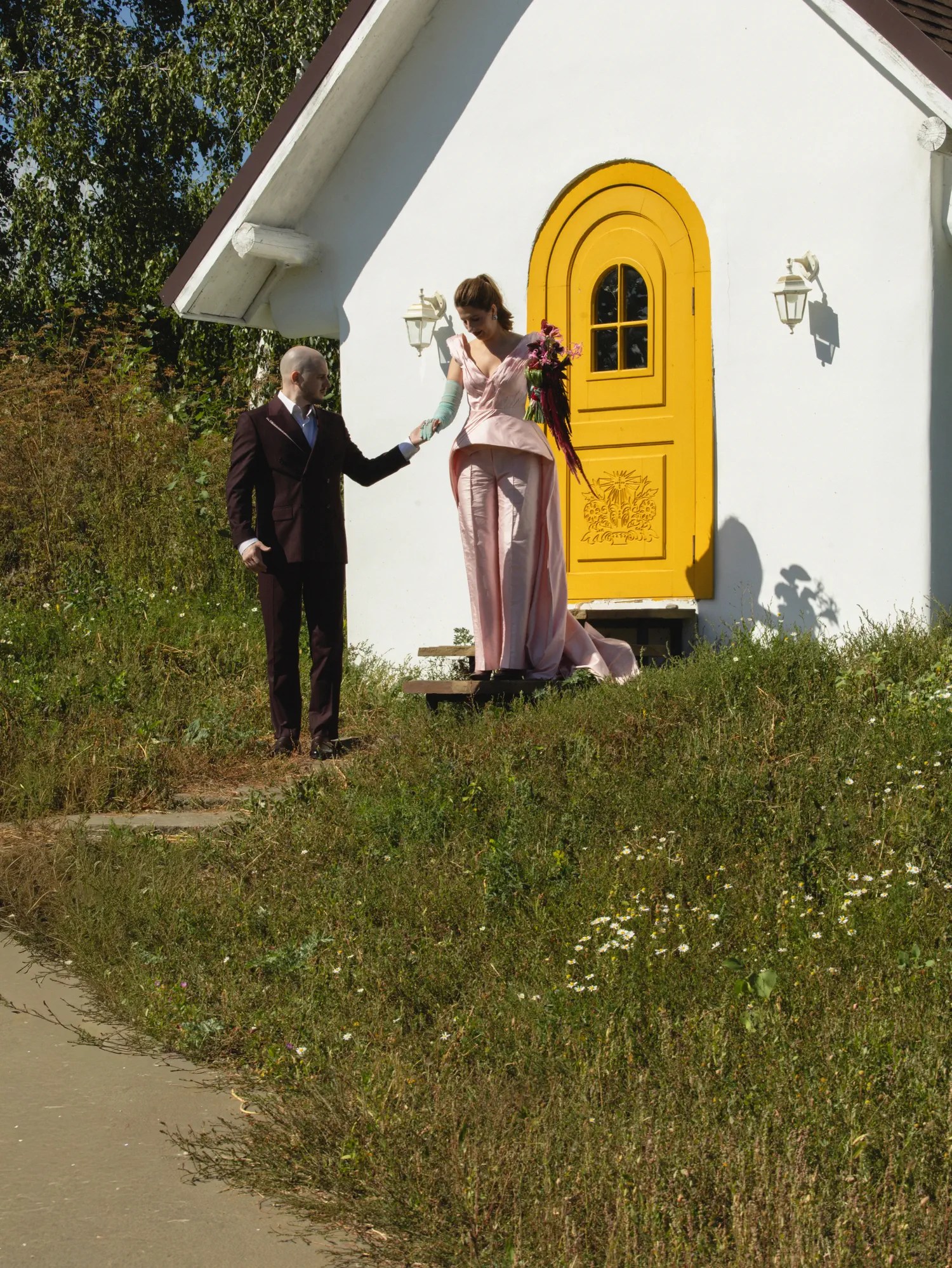 Groom reaching up to hold the bride's hand as she stands on the step of a small white chapel with a yellow door in a blush pink train gown