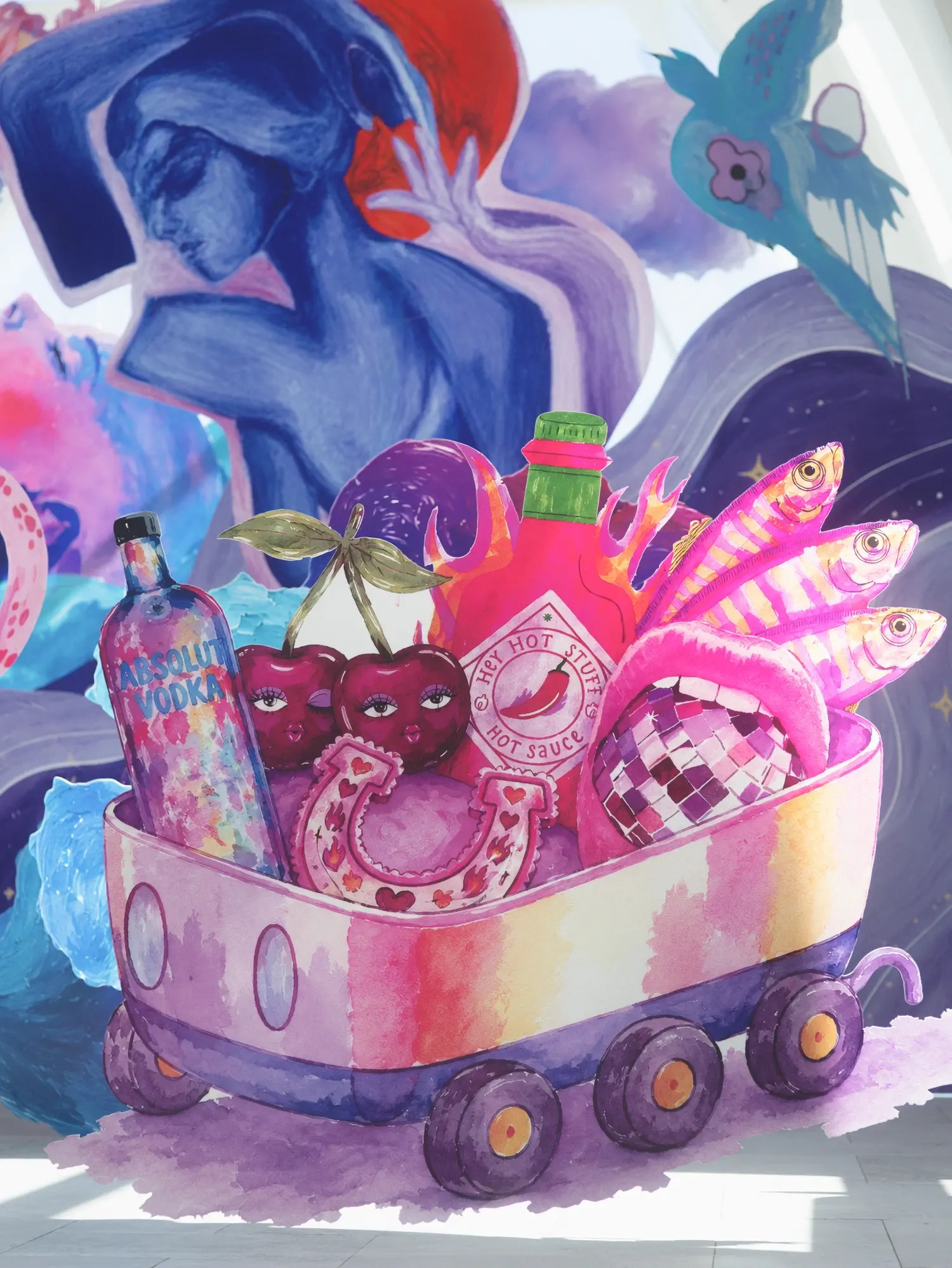 Painted panel of a rainbow toy wagon holding a vodka bottle, hot sauce, cherries with faces, a pink disco ball and striped fish