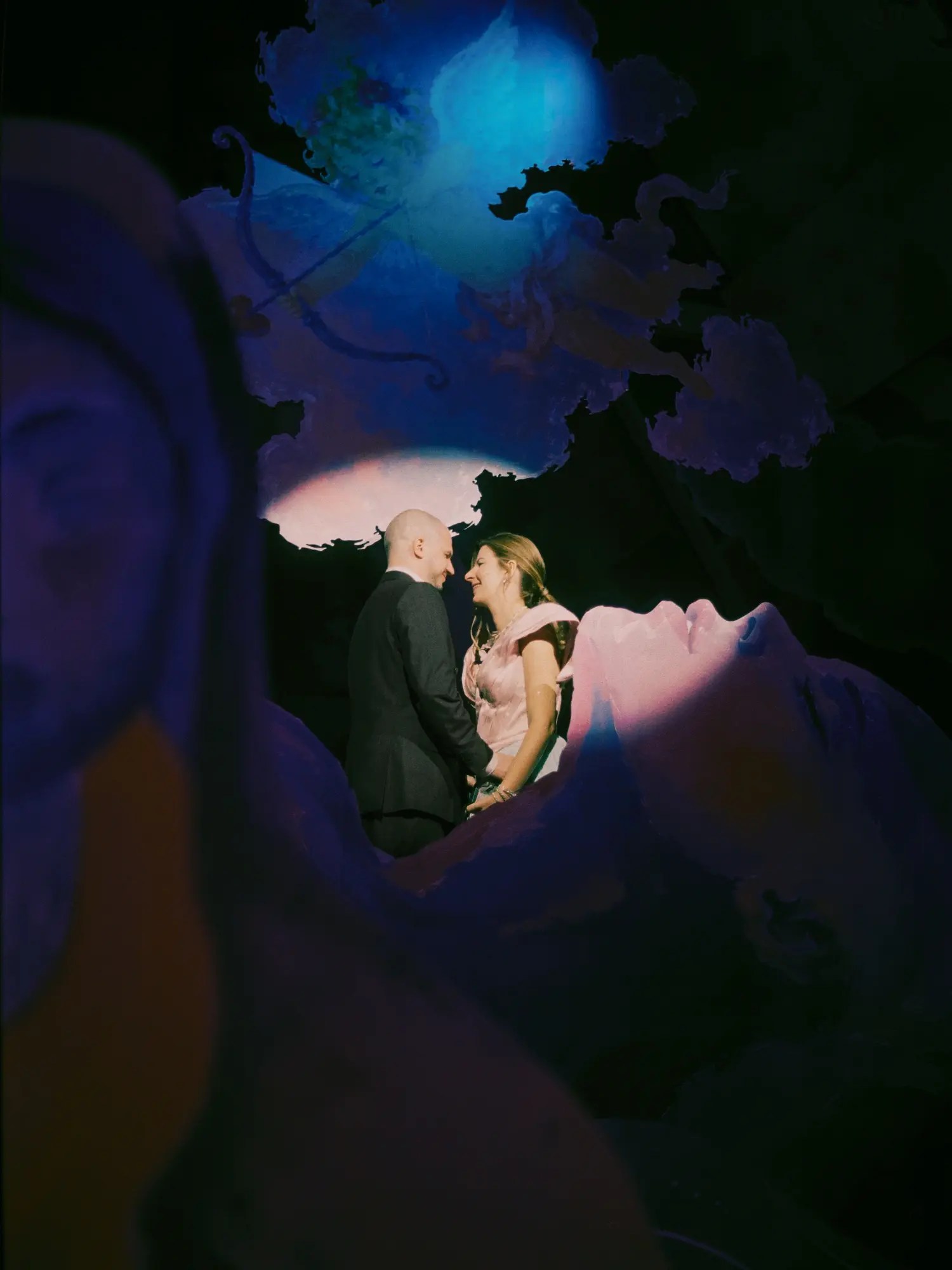Bride and groom smiling in a pool of pink light, framed through a darkened painted mural face at night
