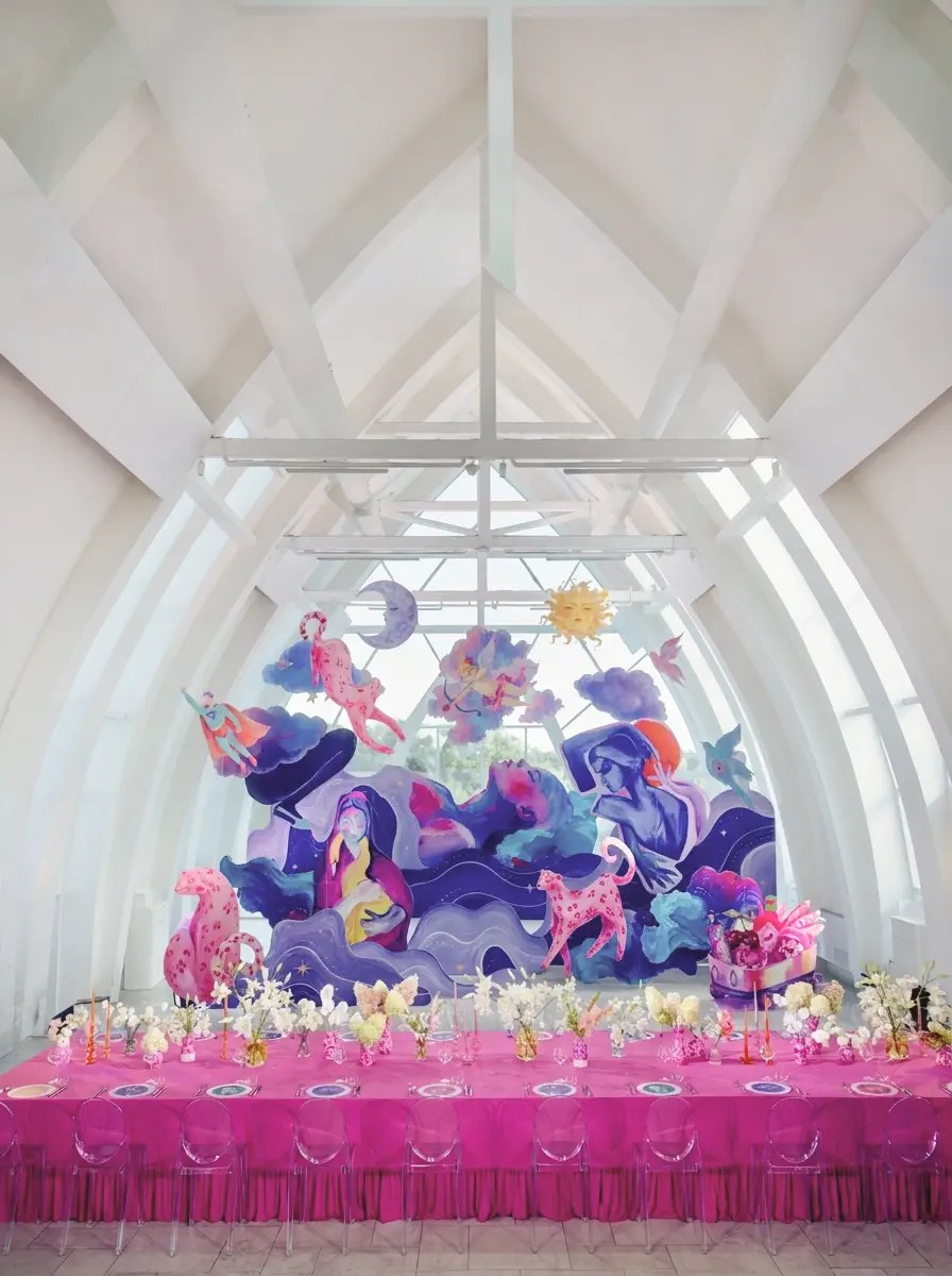 Long hot pink ruffled banquet table with clear ghost chairs before a floor-to-ceiling painted mural under a vaulted glass ceiling at Soma Island