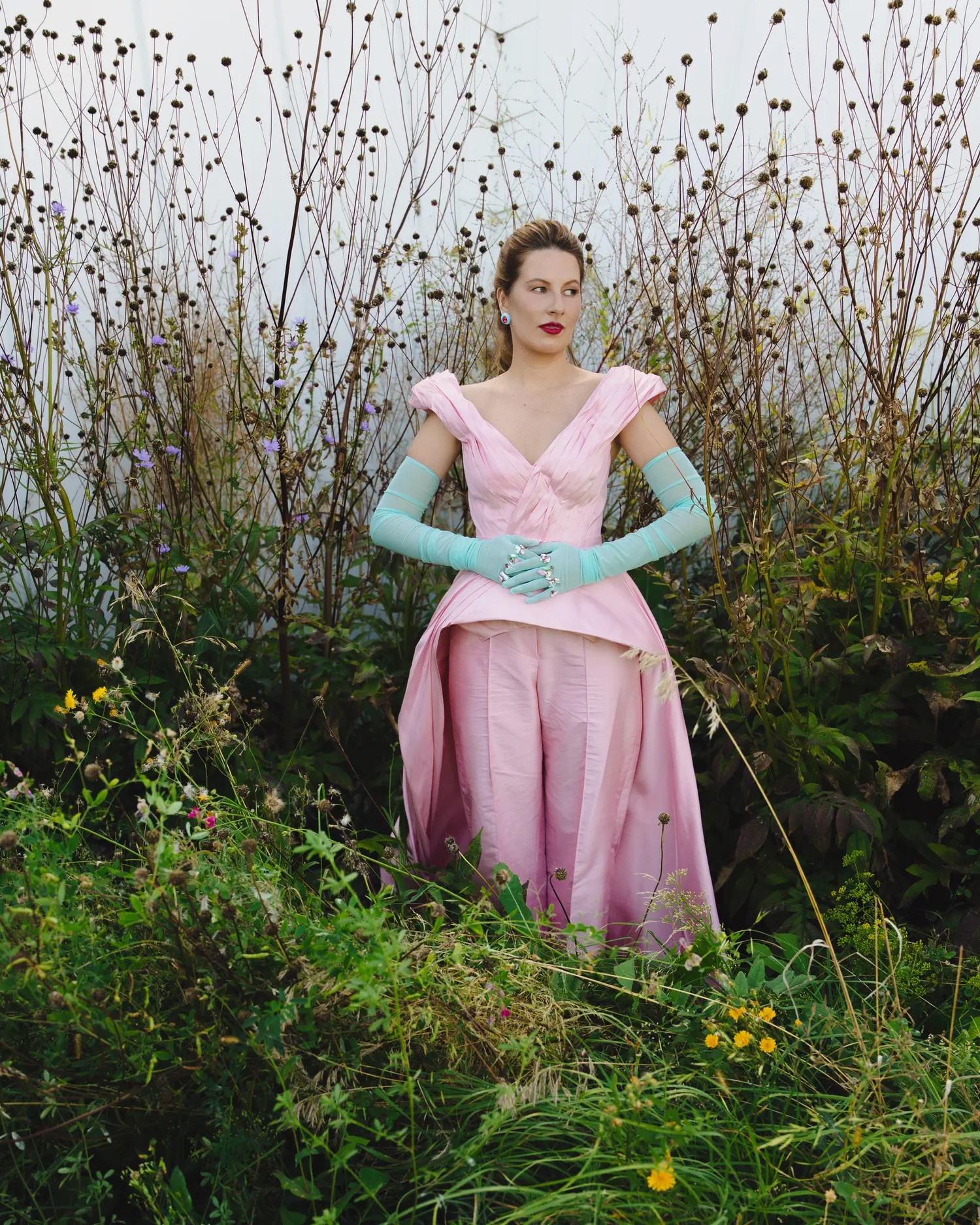 Bride in blush pink off-shoulder ball gown with pale blue sheer gloves dotted with rhinestones and a red lip standing among dried seedpod stalks