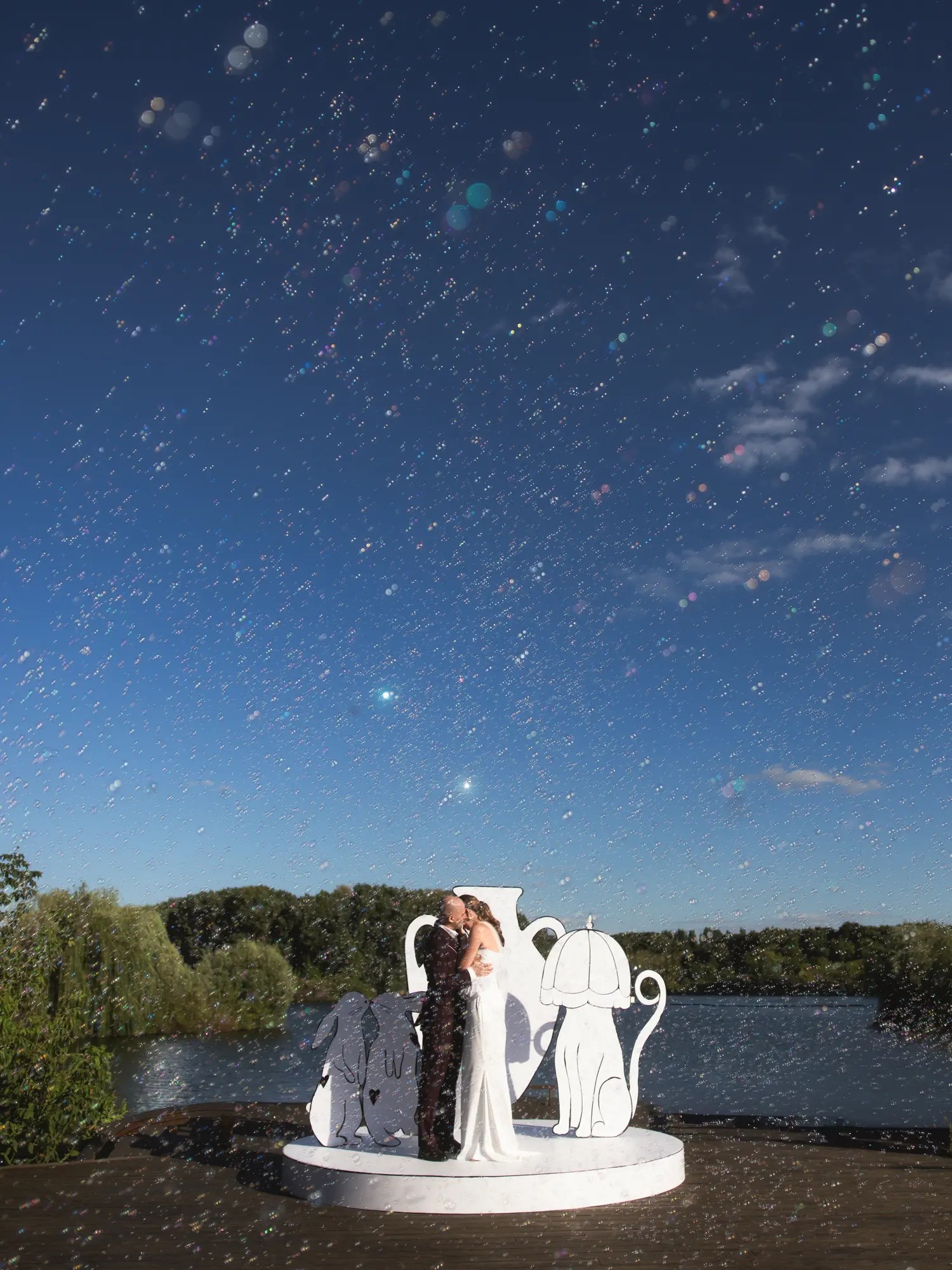 Newlyweds kissing on a lakeside ceremony platform surrounded by hundreds of soap bubbles against a blue sky