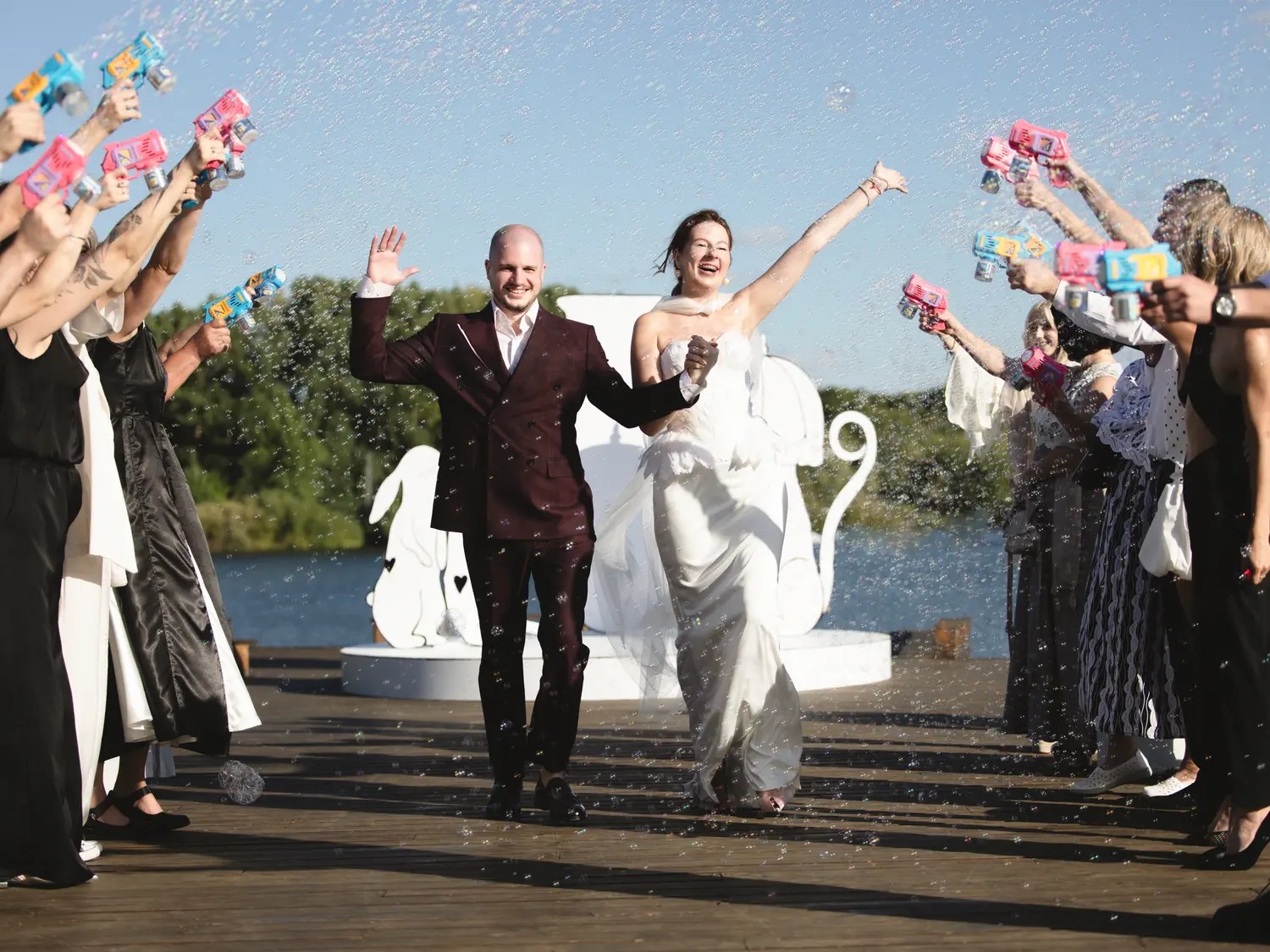 Laughing bride and groom walking a bubble-gun recessional through guests on a waterfront deck