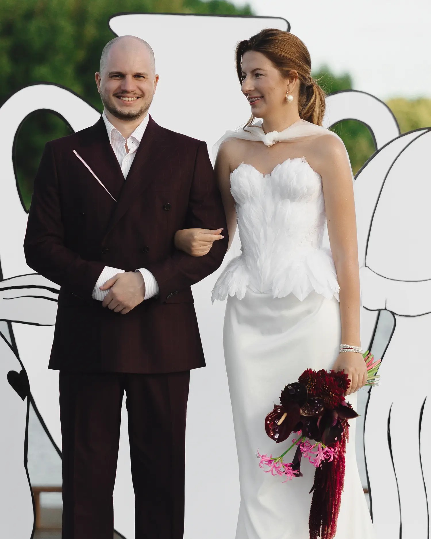Couple at the altar, bride holding a dark burgundy calla lily and pink nerine bouquet, groom in a burgundy double-breasted suit