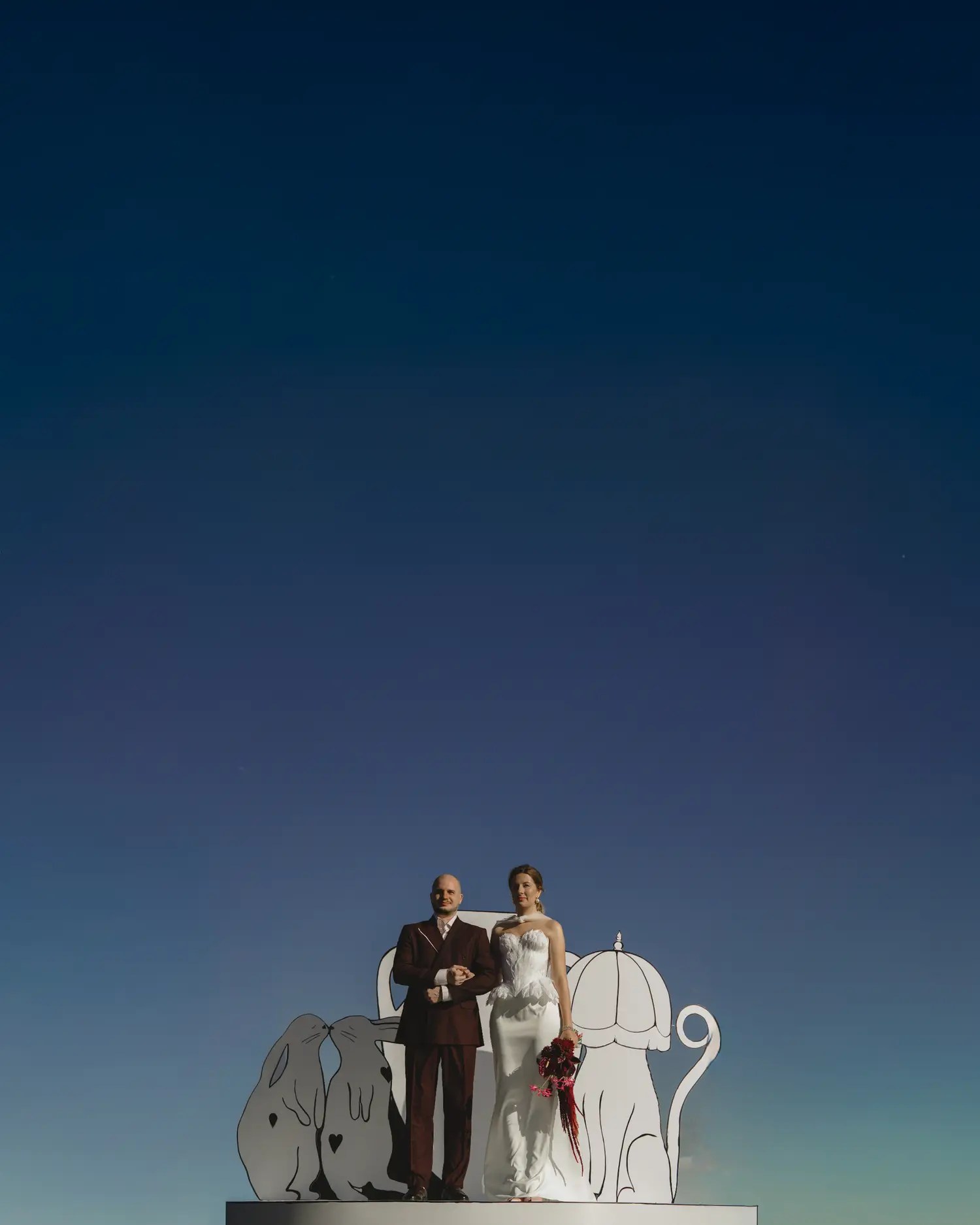 Bride in strapless white petal-appliqué gown and groom in burgundy suit on a white ceremony platform before white rabbit and cat cut-outs against a dusk sky