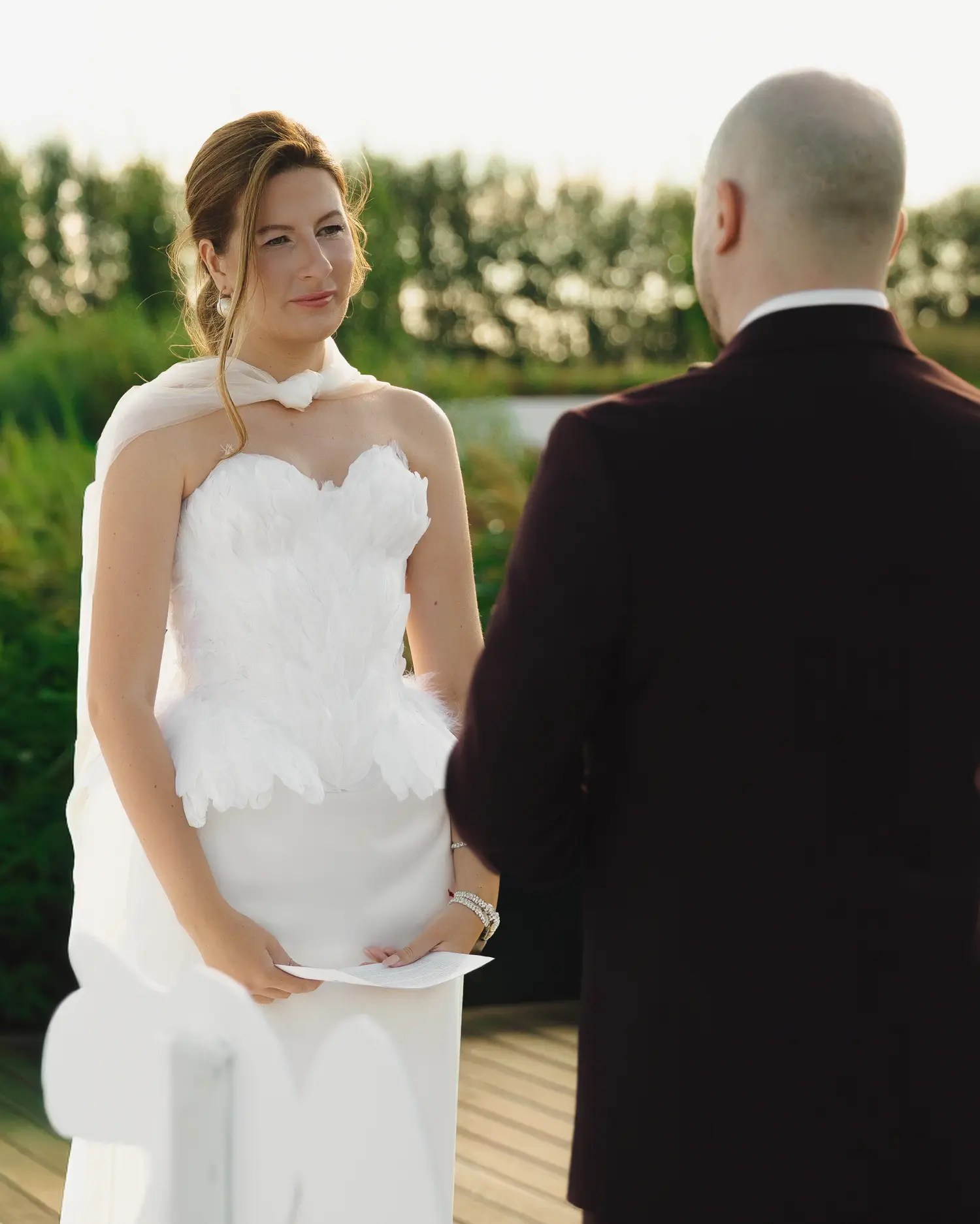 Bride with auburn updo in a white strapless gown with layered petal bodice reading vows at a golden hour lakeside ceremony