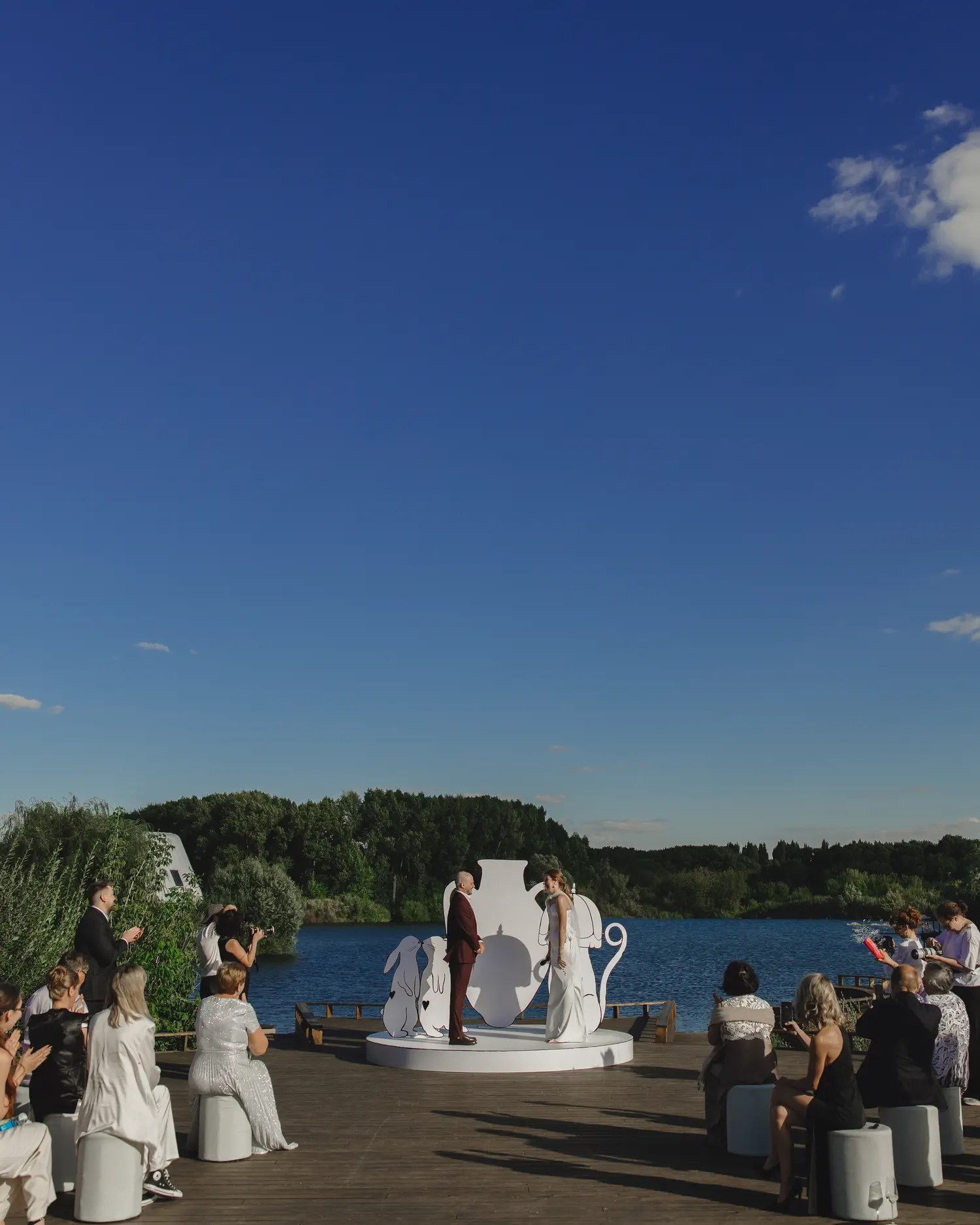 Wide view of a lakeside wedding ceremony at Soma Island with guests on white stools and a white outline art installation behind the couple