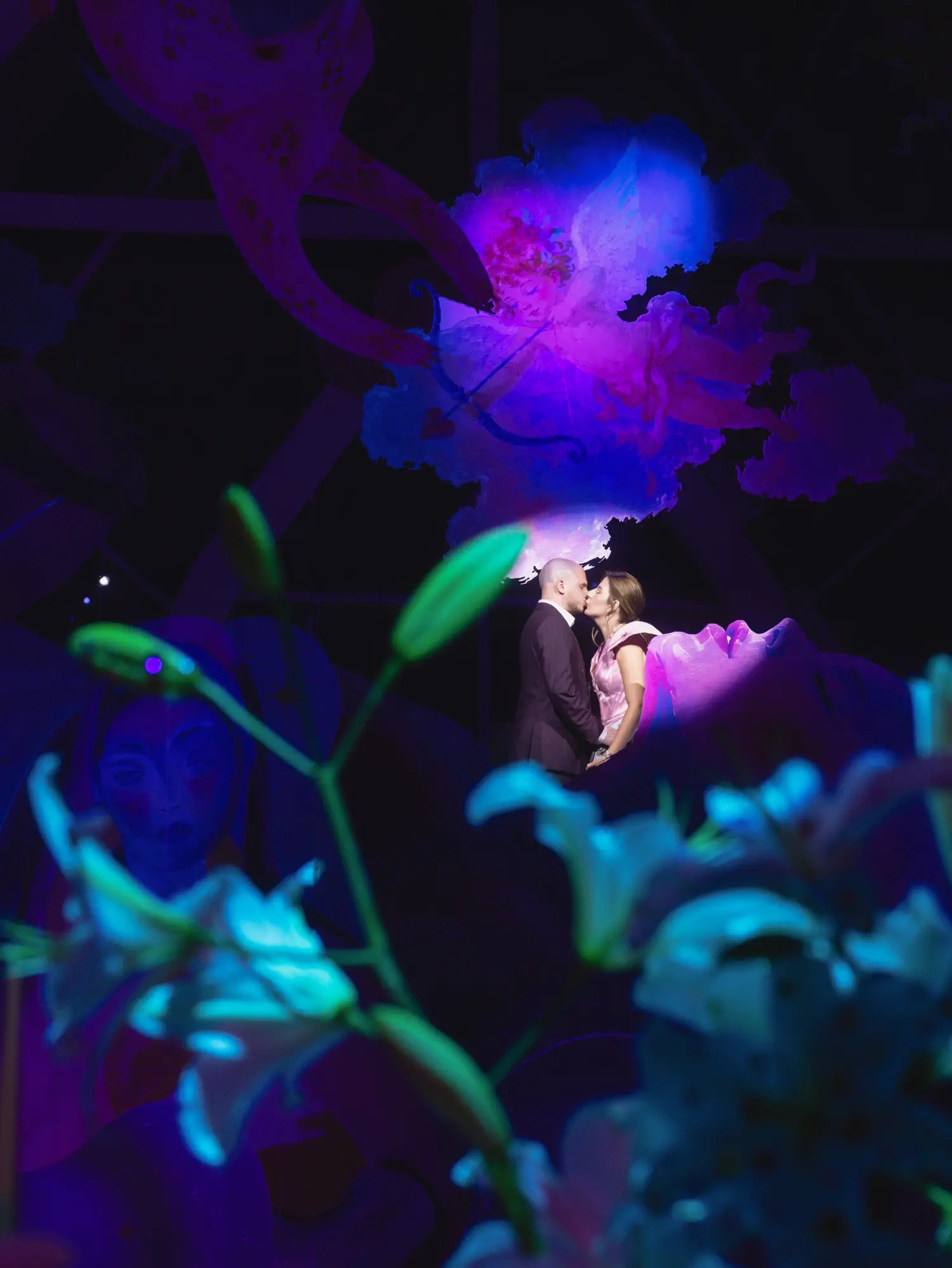 Couple kissing at night framed by lilies glowing teal under colored light with an illuminated painted backdrop
