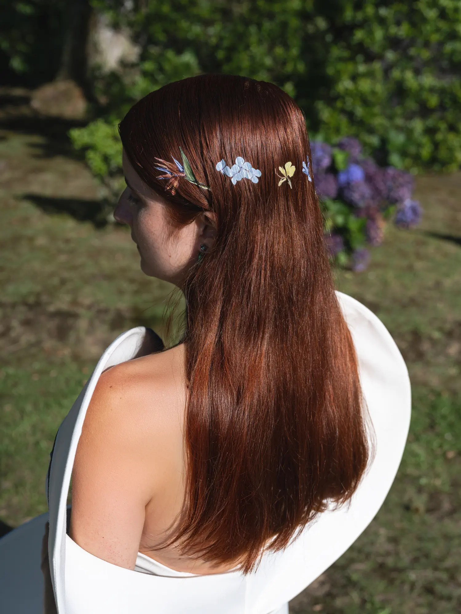 Close-up of a row of small painted botanical hair ornaments in loose auburn hair, floral motifs designed by the groom, blue hydrangeas behind
