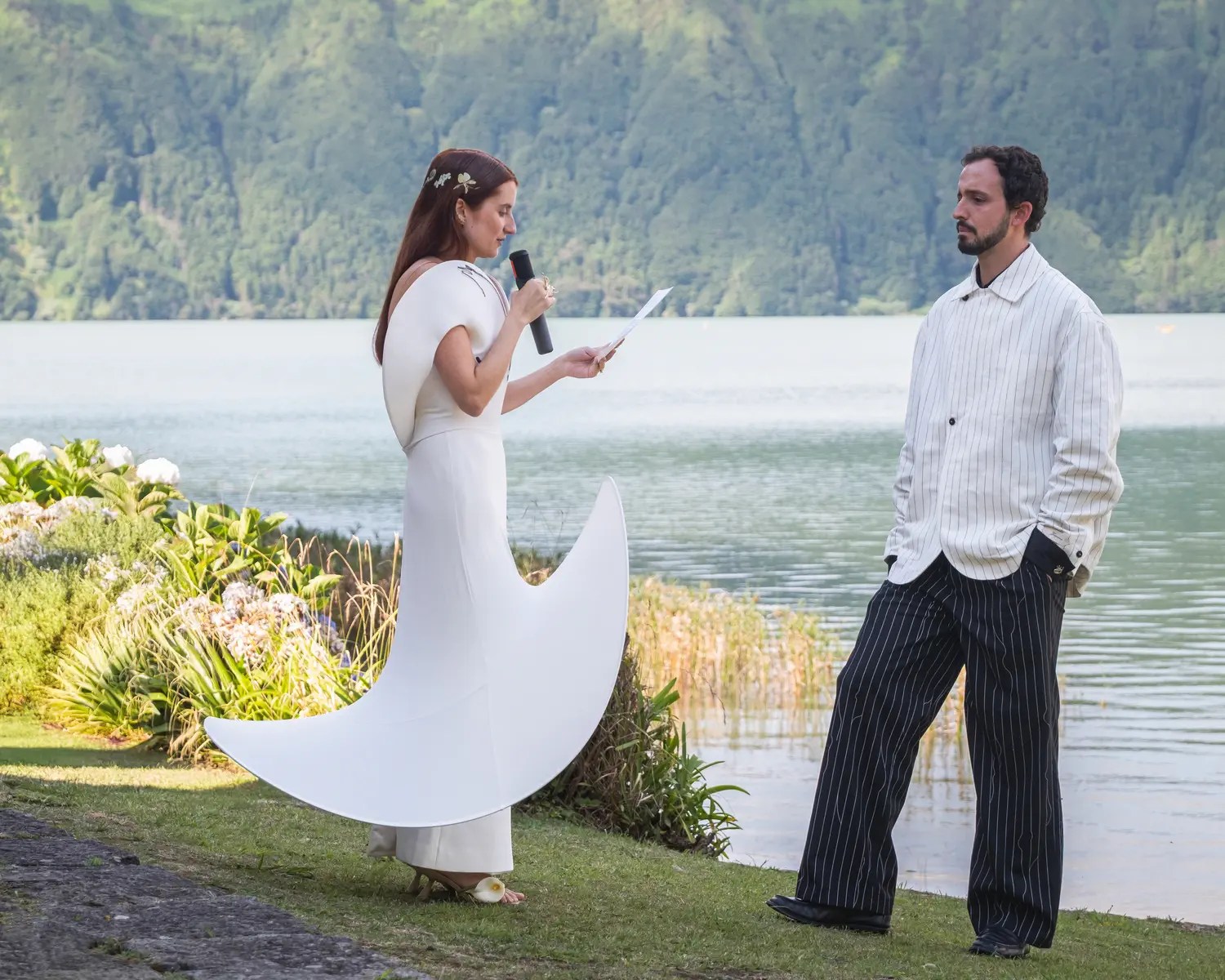Bride reading vows into a handheld microphone beside the Sete Cidades lagoon in a structural white gown and sculptural heeled sandals, groom in white pinstripe jacket and black pinstripe trousers
