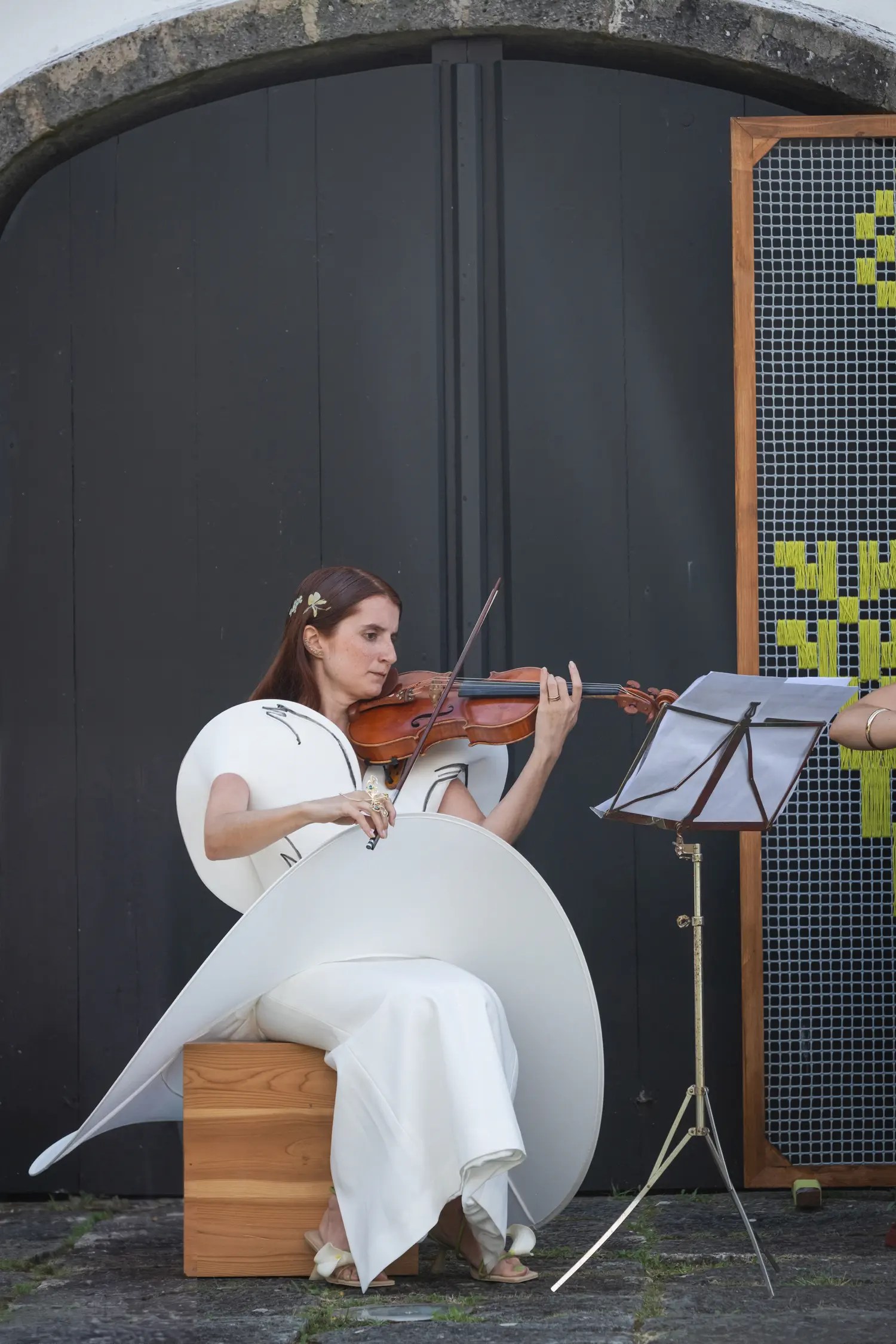 Inês playing violin in her sculptural white wedding gown, seated on a wooden block in front of a dark arched door at Casa dos Barcos
