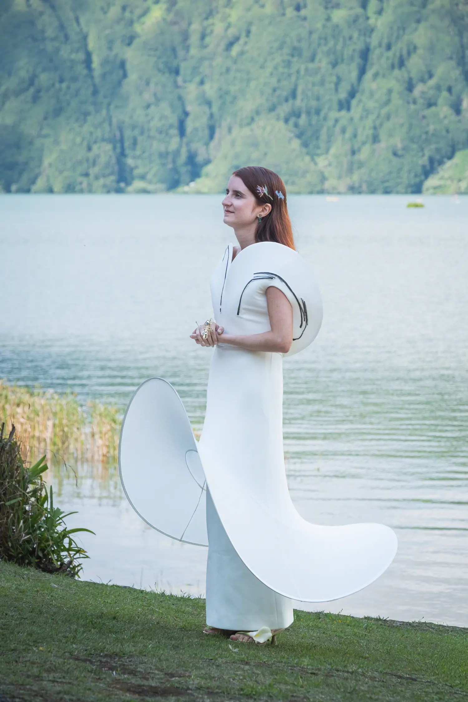 Bride in profile beside the Sete Cidades lagoon wearing a Lecia Bridal column gown with Bad Binch TONGTONG curved structural panels sweeping from shoulder to ground