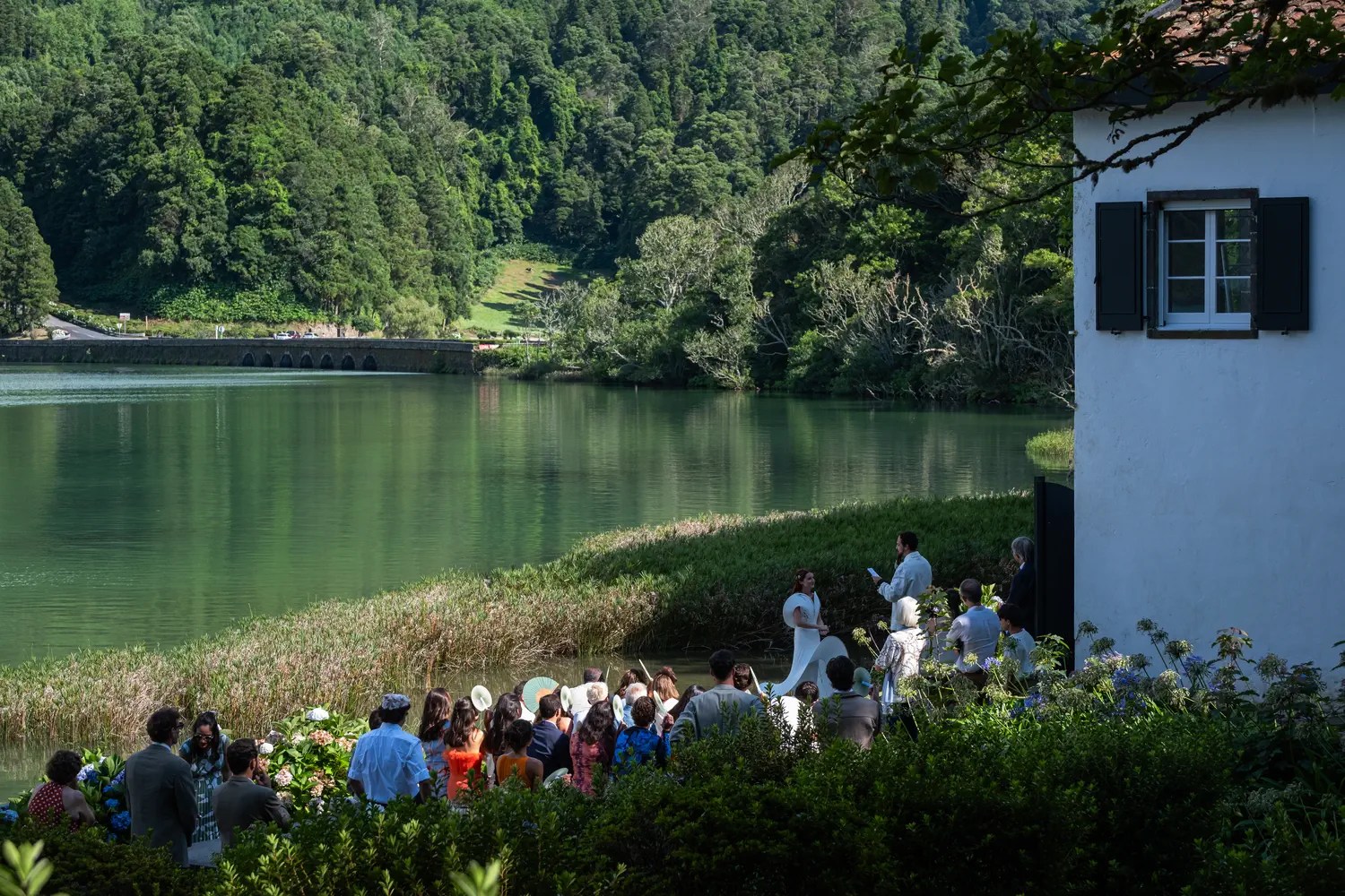 Elevated view of an intimate lakeside wedding ceremony at Sete Cidades, guests gathered on the grass with a stone arched bridge and forested caldera behind