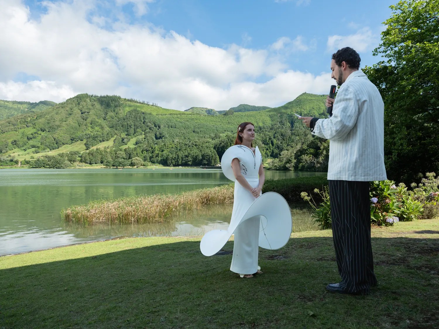 Groom reading vows to the bride on a lawn beside the Sete Cidades lagoon under blue sky, white pinstripe jacket and black pinstripe trousers, bride in sculptural white gown