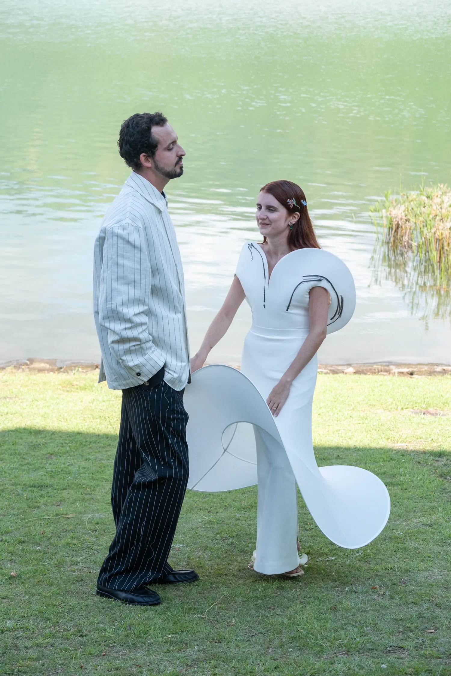 Couple holding hands and smiling at the water's edge of a green caldera lake in São Miguel, bride in sculptural white gown with floral hair ornaments