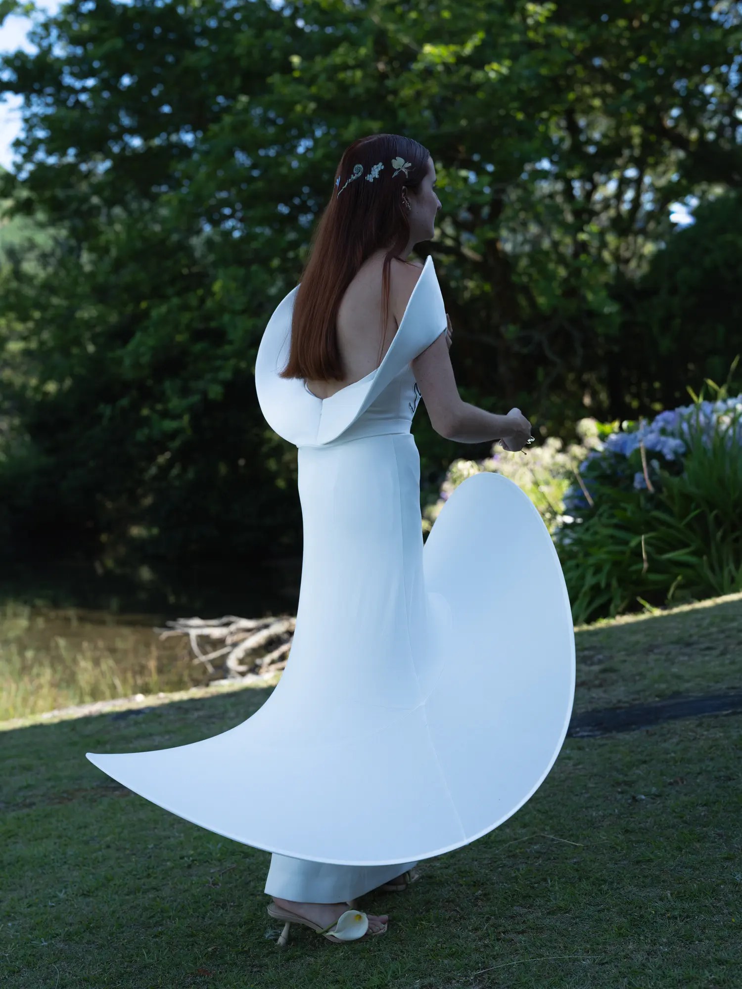Back view of bride's open-back sculptural white gown with trailing curved panel and painted floral hair ornaments in a shaded Azores garden with agapanthus