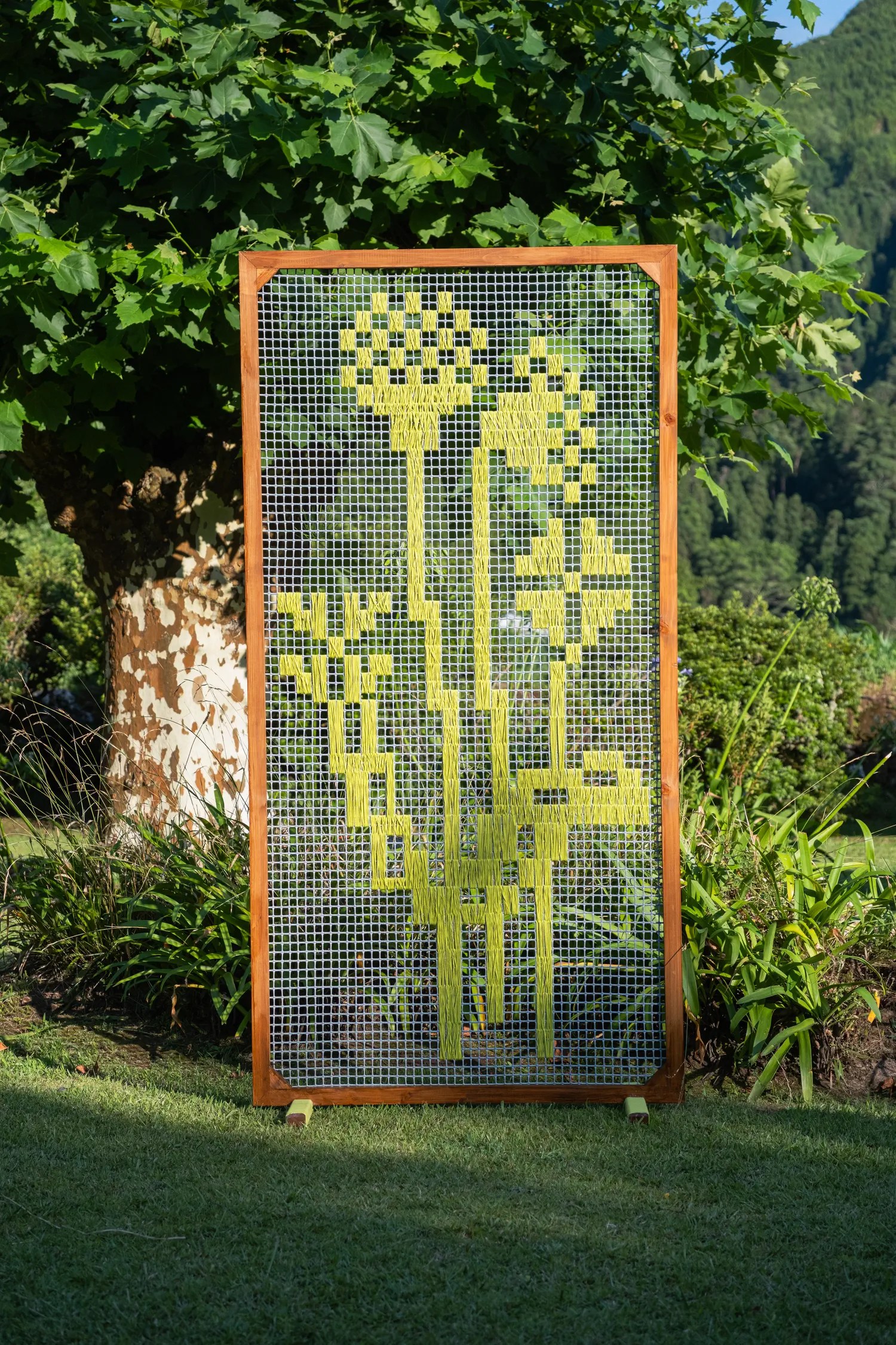 Freestanding wooden-framed grid panel woven with lime green yarn into large block letters on a garden lawn in the Azores