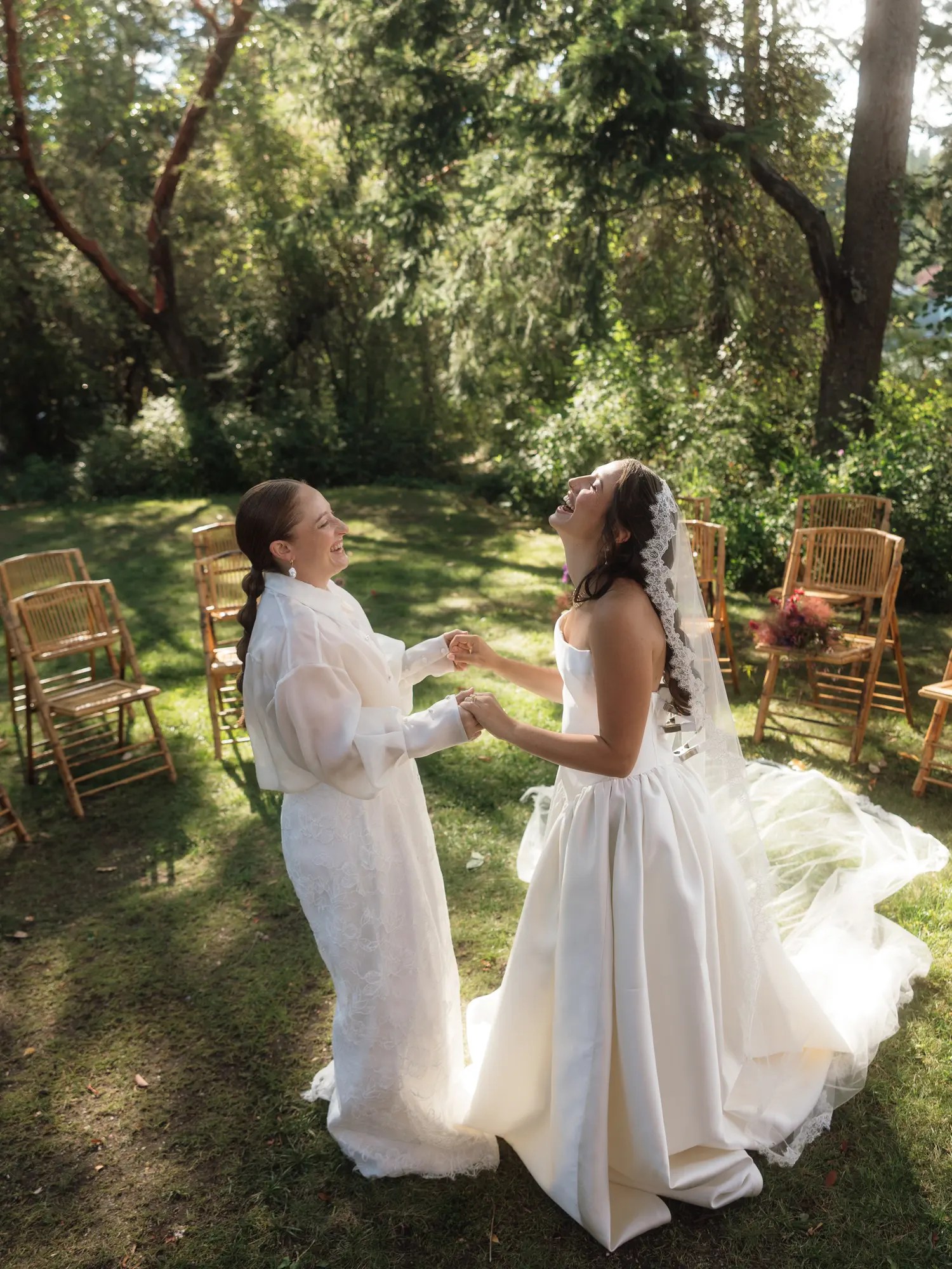 Two brides laughing and spinning on a sunlit lawn, one in a strapless ivory gown with lace veil and the other in an organza barong with lace wide-leg trousers