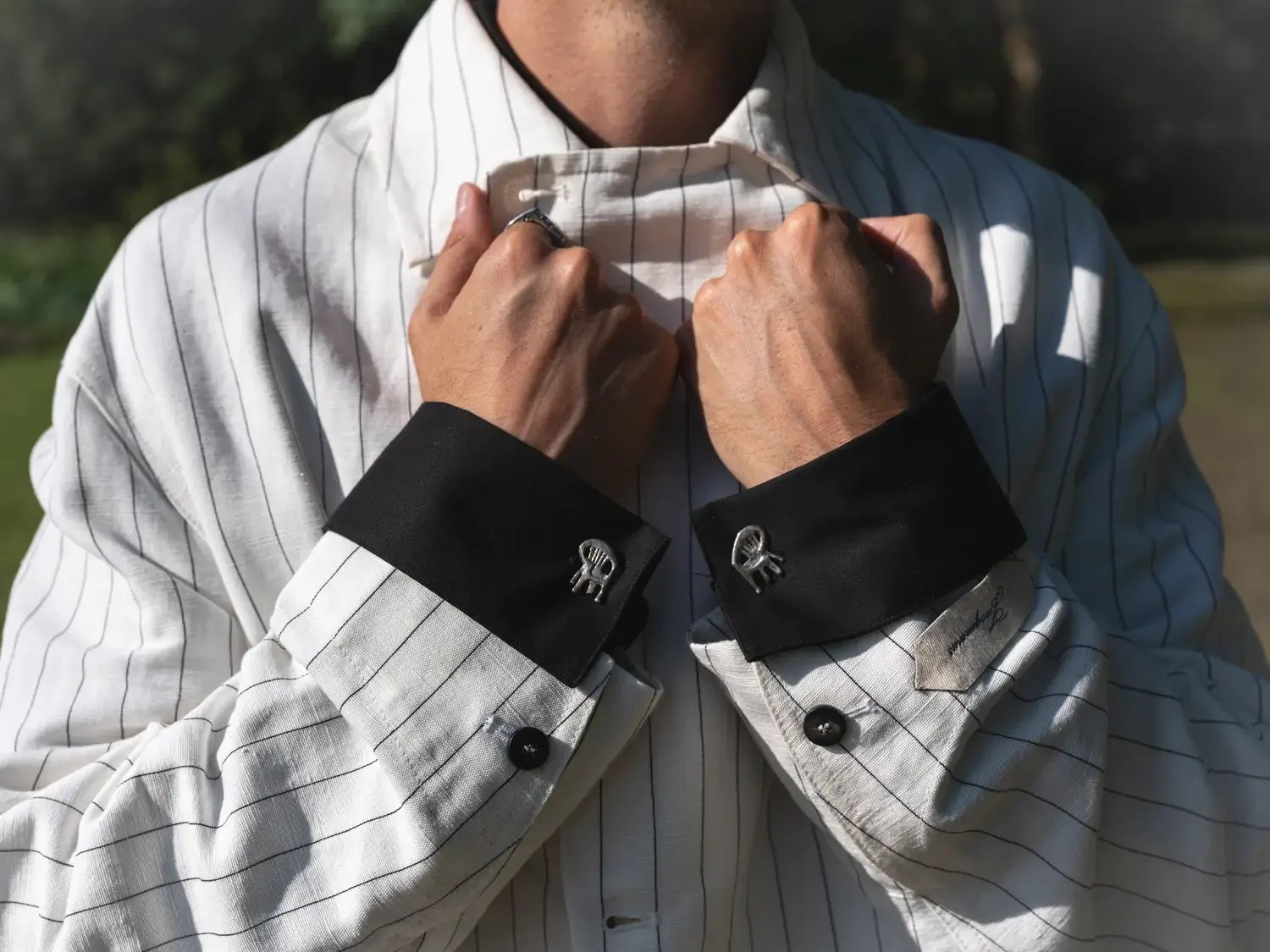 Close-up of the groom adjusting the collar of a white pinstripe Jacquemus jacket with black cuffs, fastened with silver chair-shaped cufflinks