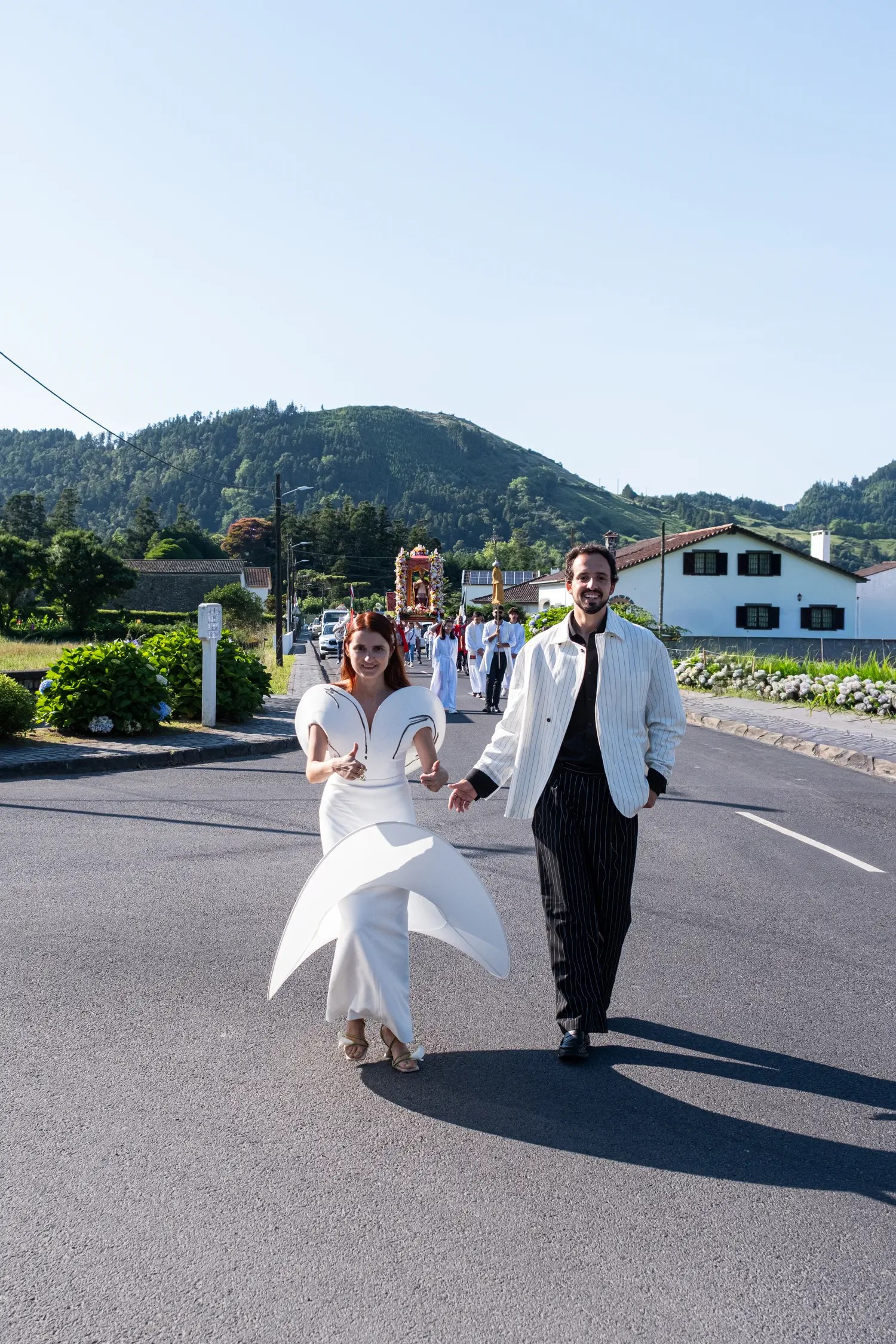 Bride in a sculptural white gown and groom in a white pinstripe jacket walking hand in hand down a road in Sete Cidades, a local religious procession following behind