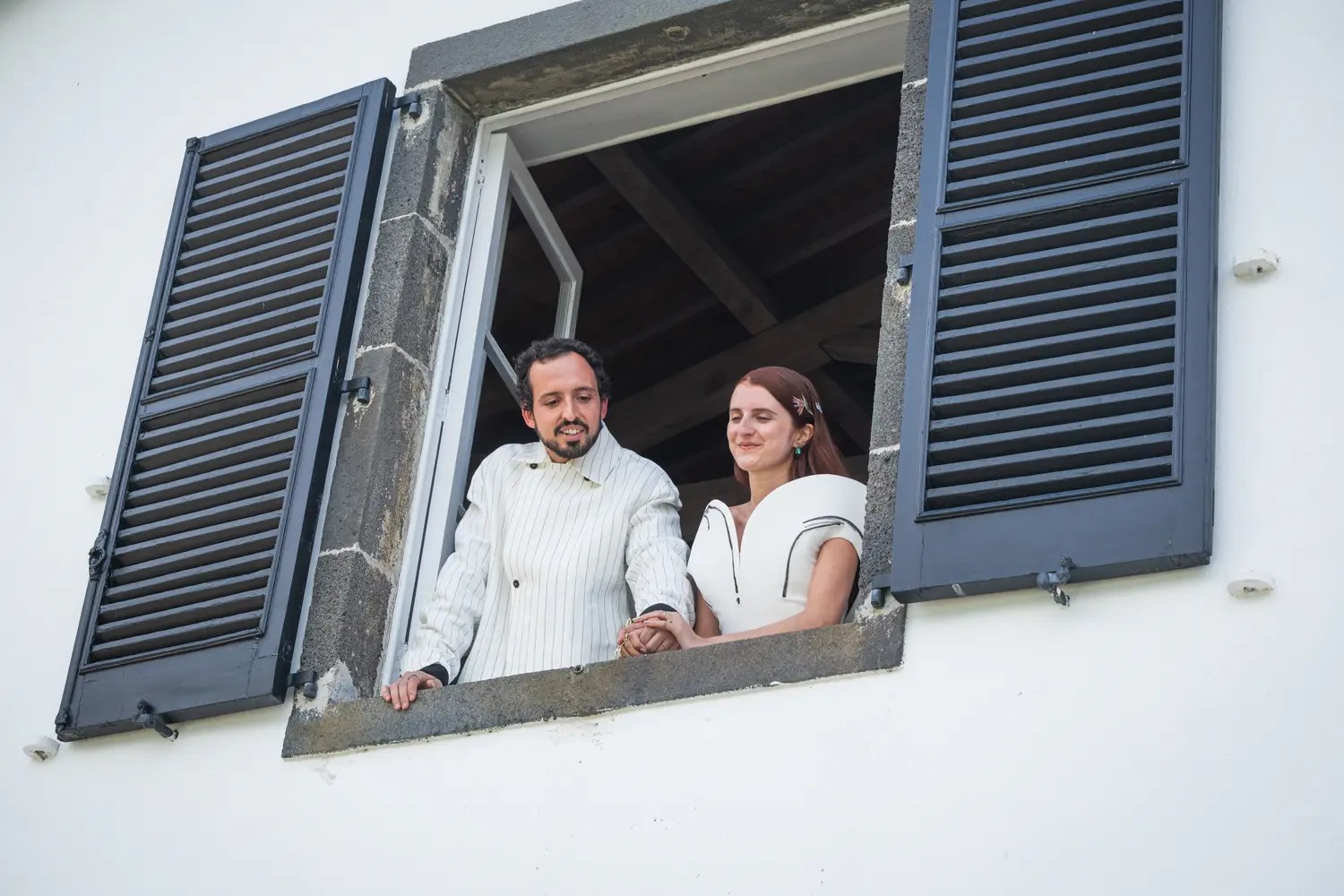Inês and Rodrigo looking down from the upper window of the Casa dos Barcos boat house on the Sete Cidades lagoon, framed by navy shutters on a whitewashed wall