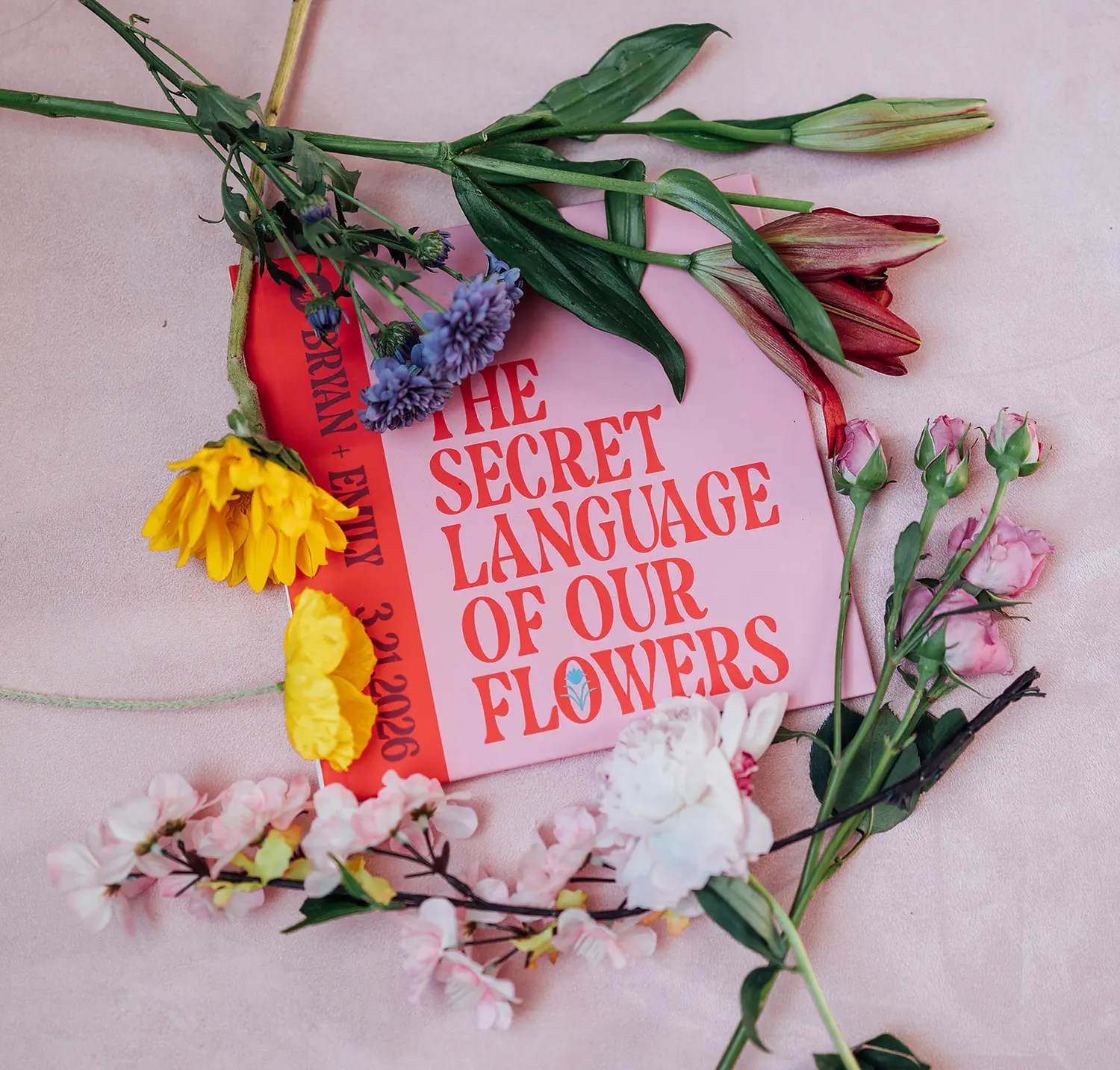 Red booklet titled The Secret Language of Our Flowers surrounded by loose lily, thistle, daisy, and sweet pea blooms on blush fabric