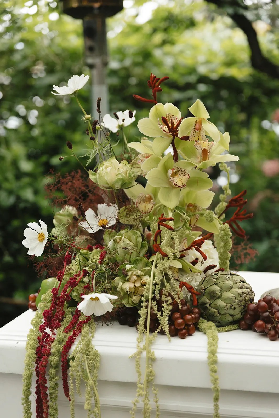 Arrangement de cérémonie inspiré du jardin composé d'orchidées cymbidium vertes, d'artichauts, de cosmos blanc, de patte de kangourou rouge, d'amarante et de raisins noirs