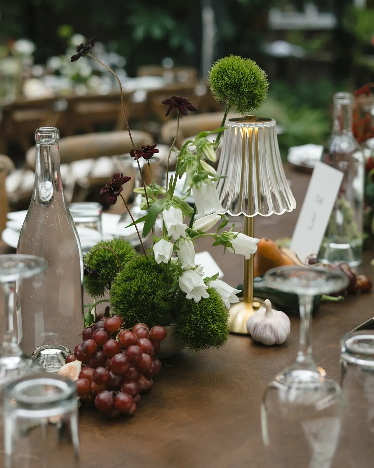 Détail de la table de réception avec lampe en laiton, globe d'allium vert, cosmos sombre, fleurs en cloche blanches, raisins rouges, bulbe d'ail et tranches de figue sur noyer