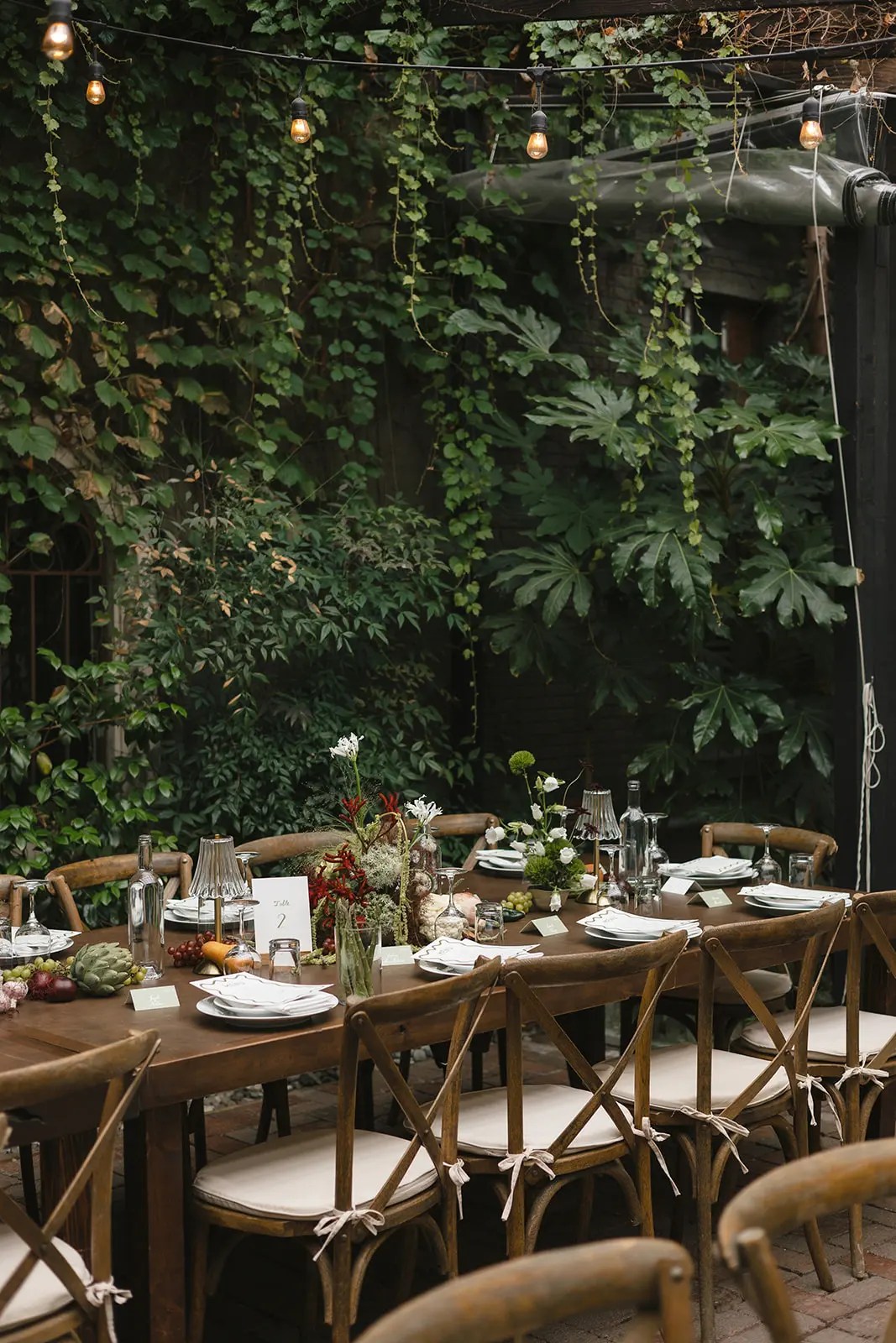 Longue table de ferme en noyer foncé avec chaises à dossier croisé et centres de table sous des guirlandes lumineuses Edison contre un mur de feuillage vert au Corson Building