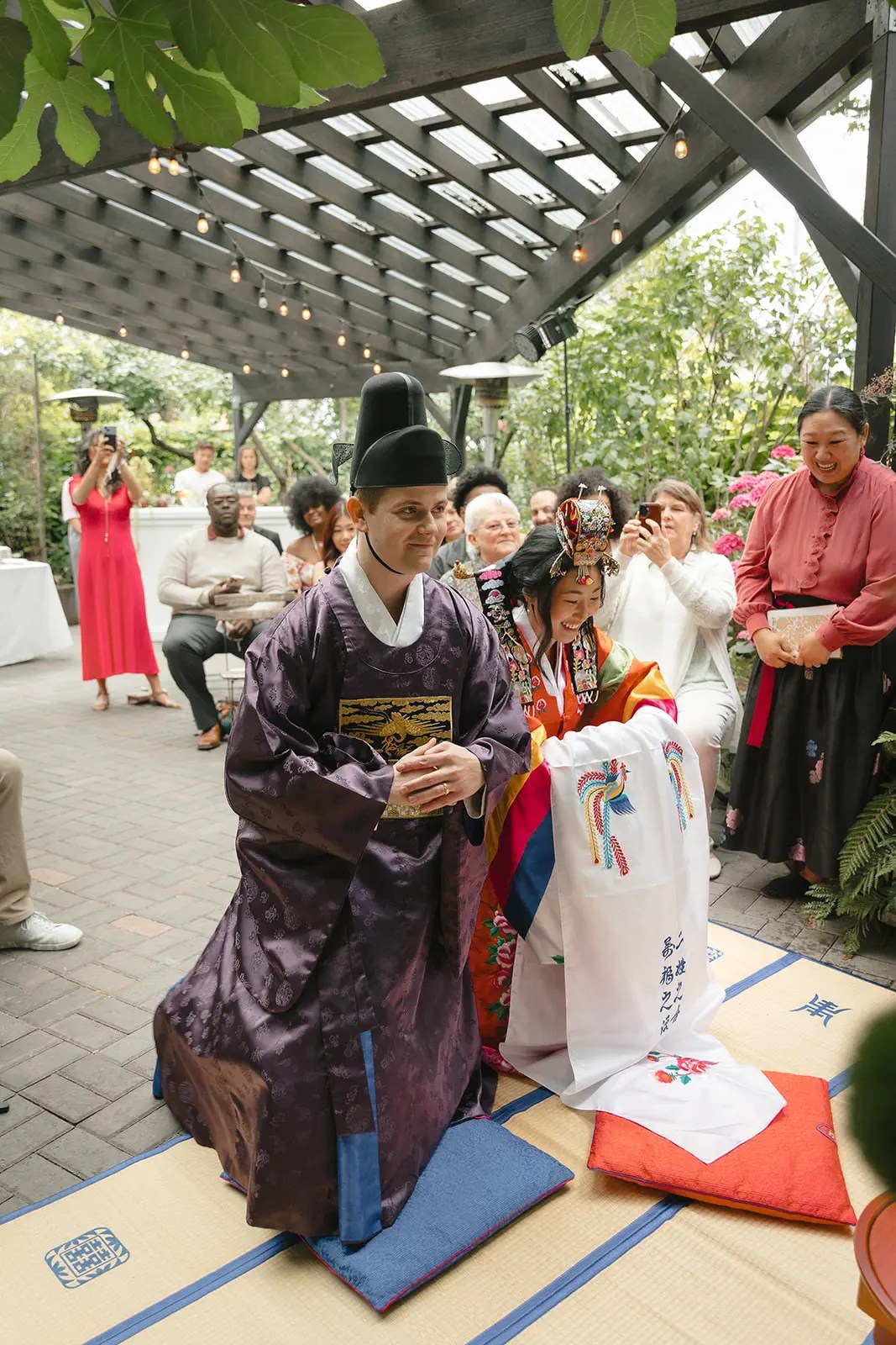 Couple s'agenouillant et s'inclinant sur des nattes tissées dans un hanbok traditionnel lors d'une cérémonie coréenne Paebaek sous une pergola de jardin avec des invités rassemblés à proximité