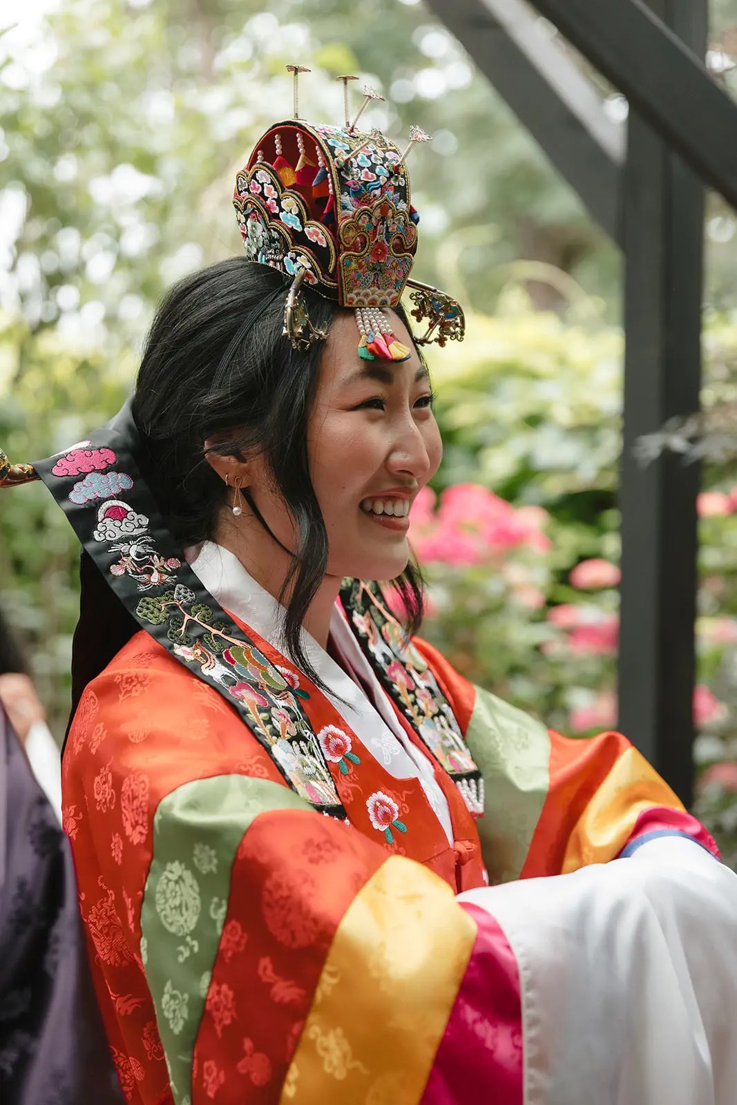 Mariée souriante en coiffure de mariée coréenne brodée avec ornements en perles et hanbok orange avec broderie phénix, hortensias roses derrière